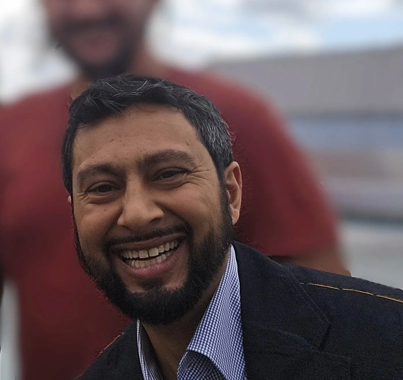 Smiling man with dark hair, beard, wearing a suit jacket and checkered shirt, in front of a blurred background with another person behind him. This is Arjun Tasker, Local Works Global co-founder and senior advisor of partnerships.