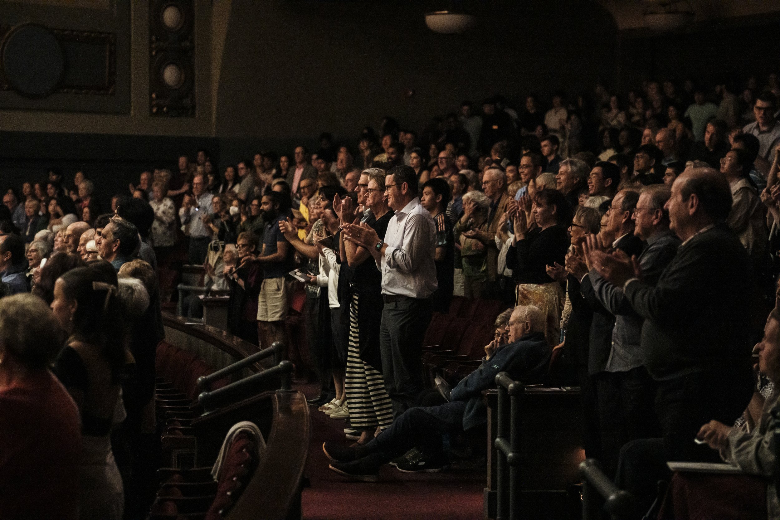 An audience gives a standing ovation after a performance in an auditorium.