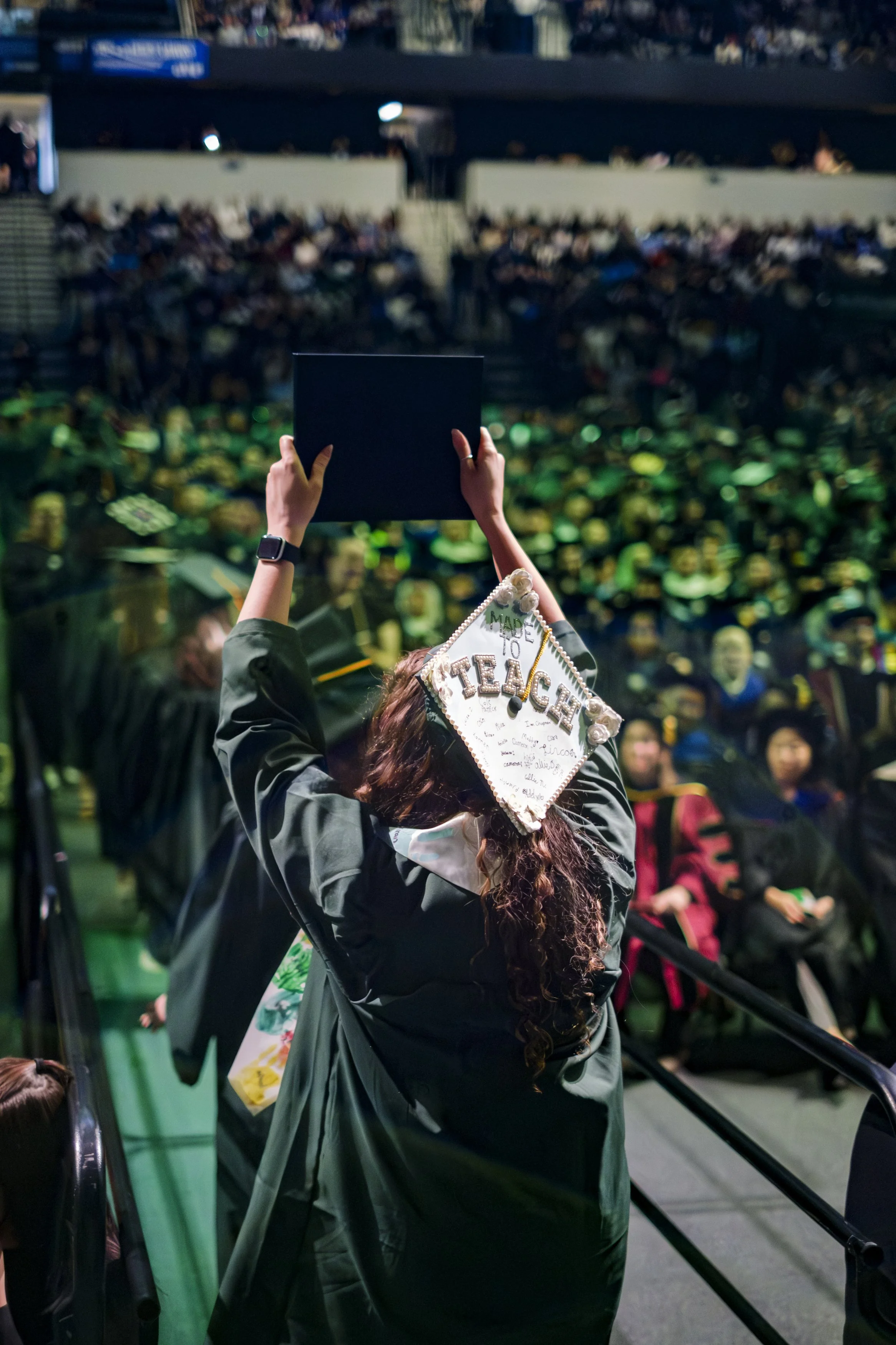 A person with a decorated graduation cap that reads "TEACH" holds up their diploma while walking toward a crowd of people..