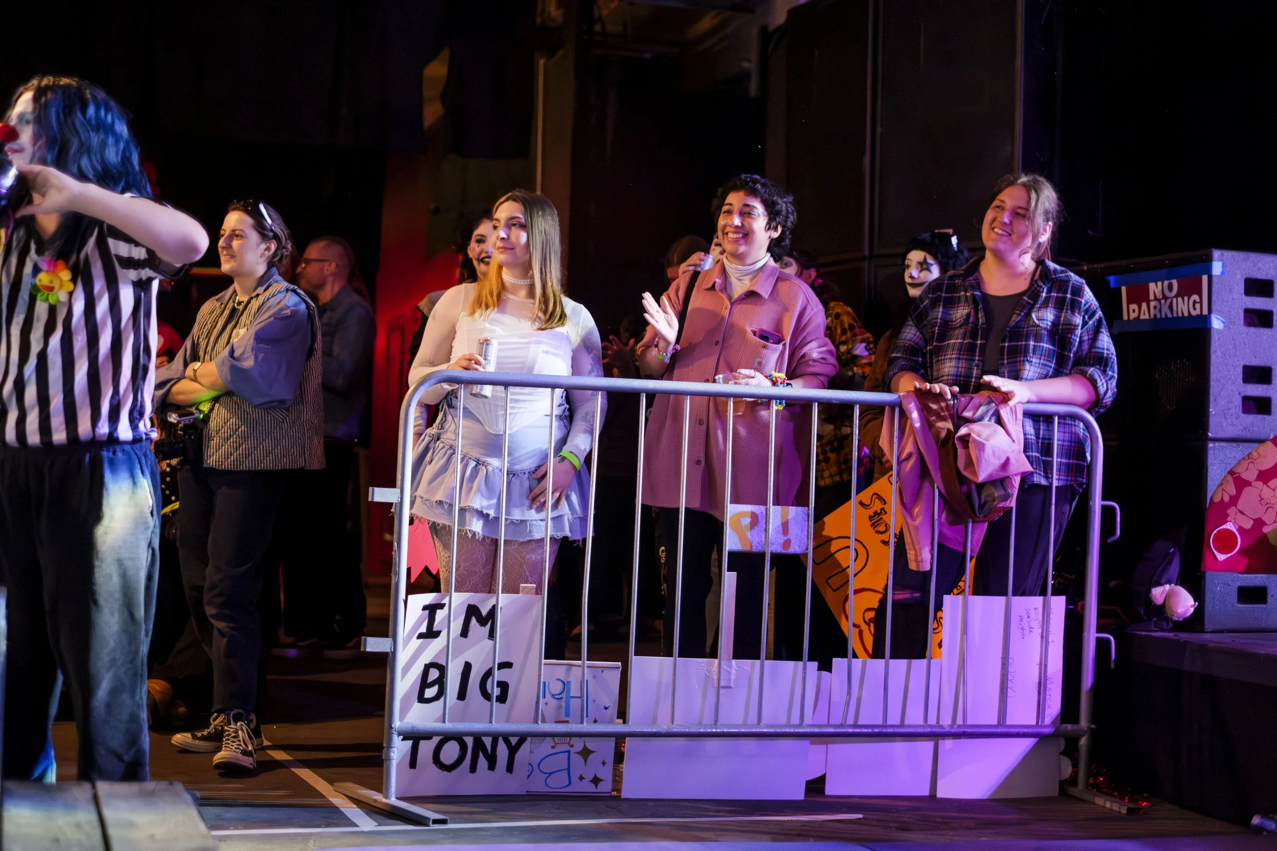 A group of people in the audience stand behind barriers and smile while clapping and holding drinks.
