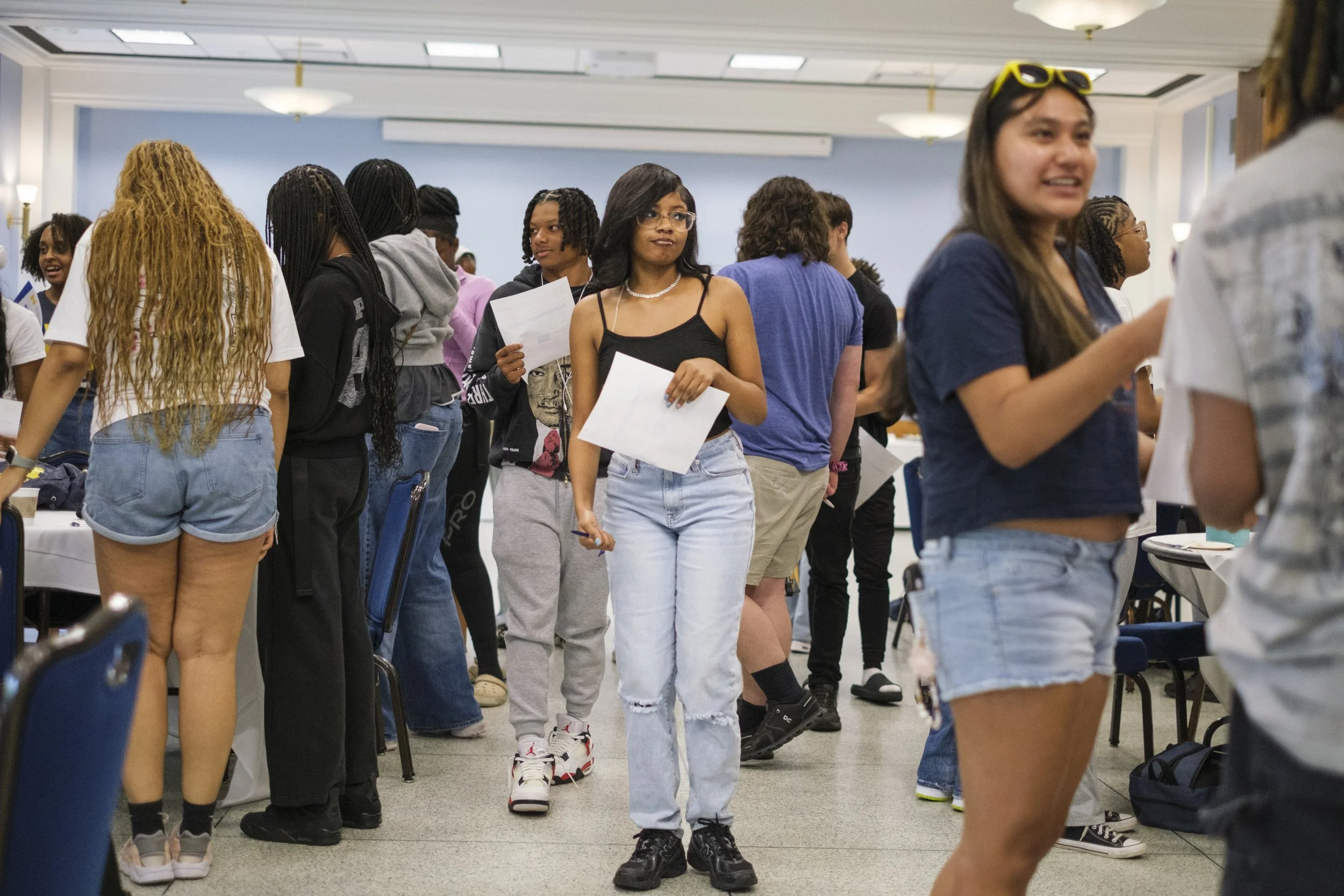 A group of high school aged students walk around a room while holding papers and introduce themselves to one another.