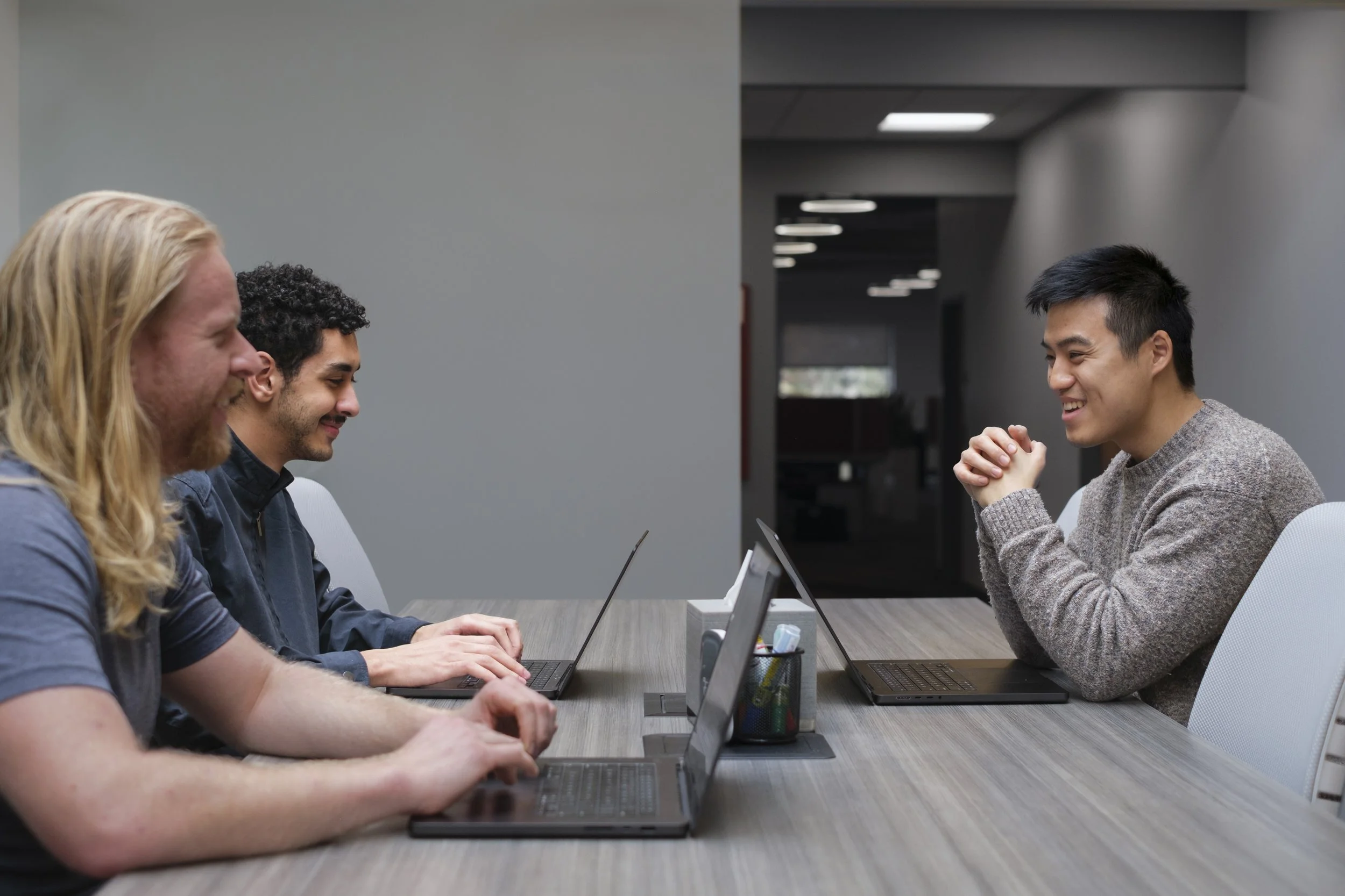 Three people with laptops open on a table top smile at one another in an office space.
