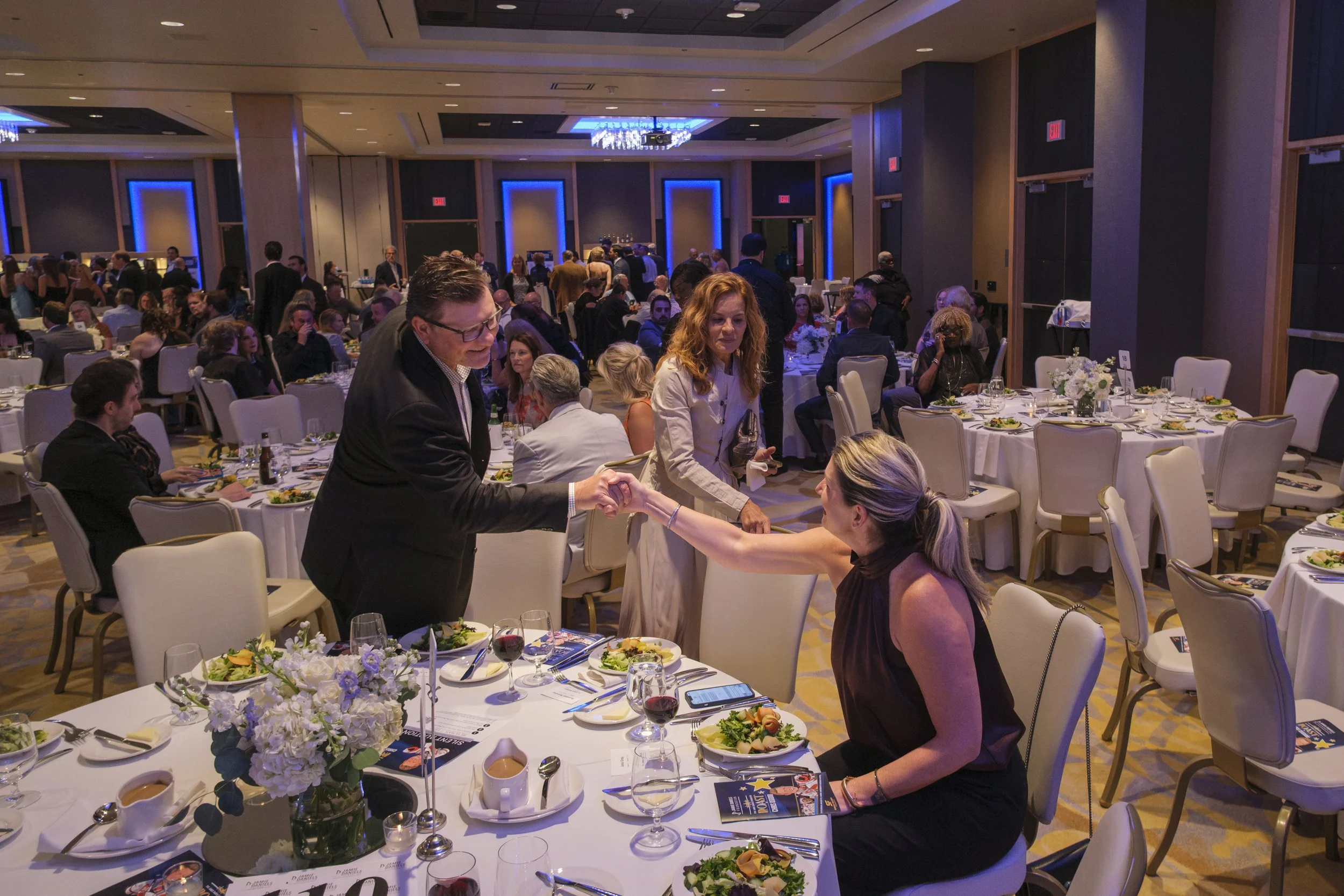 A woman seated at a table with food shakes hands with a man wearing a suit during a gala.