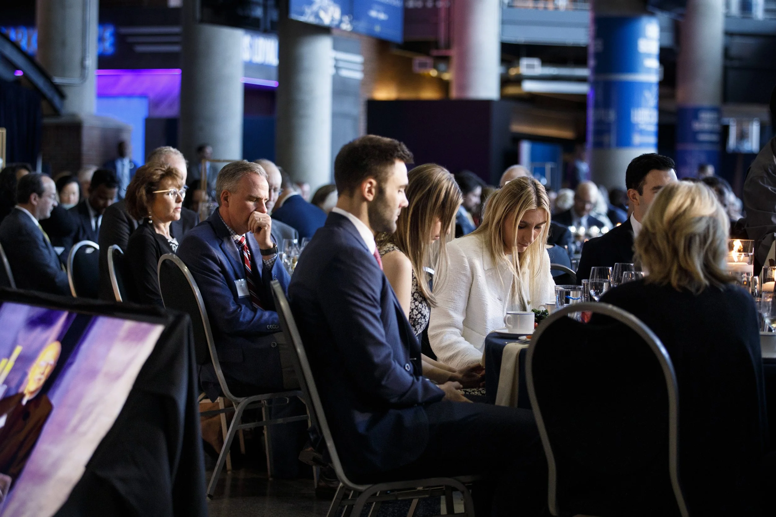 Gala guests sit at a table, bowing their heads in prayer.