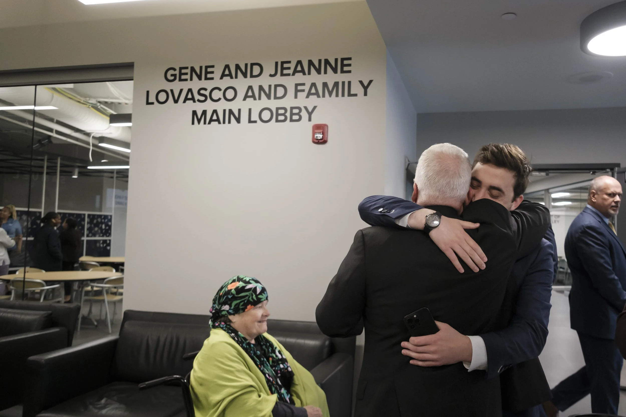 A man closes his eyes while he hugs another man. Behind them is a sign that reads "Gene and Jeanne Lovasco and Family Main Lobby".
