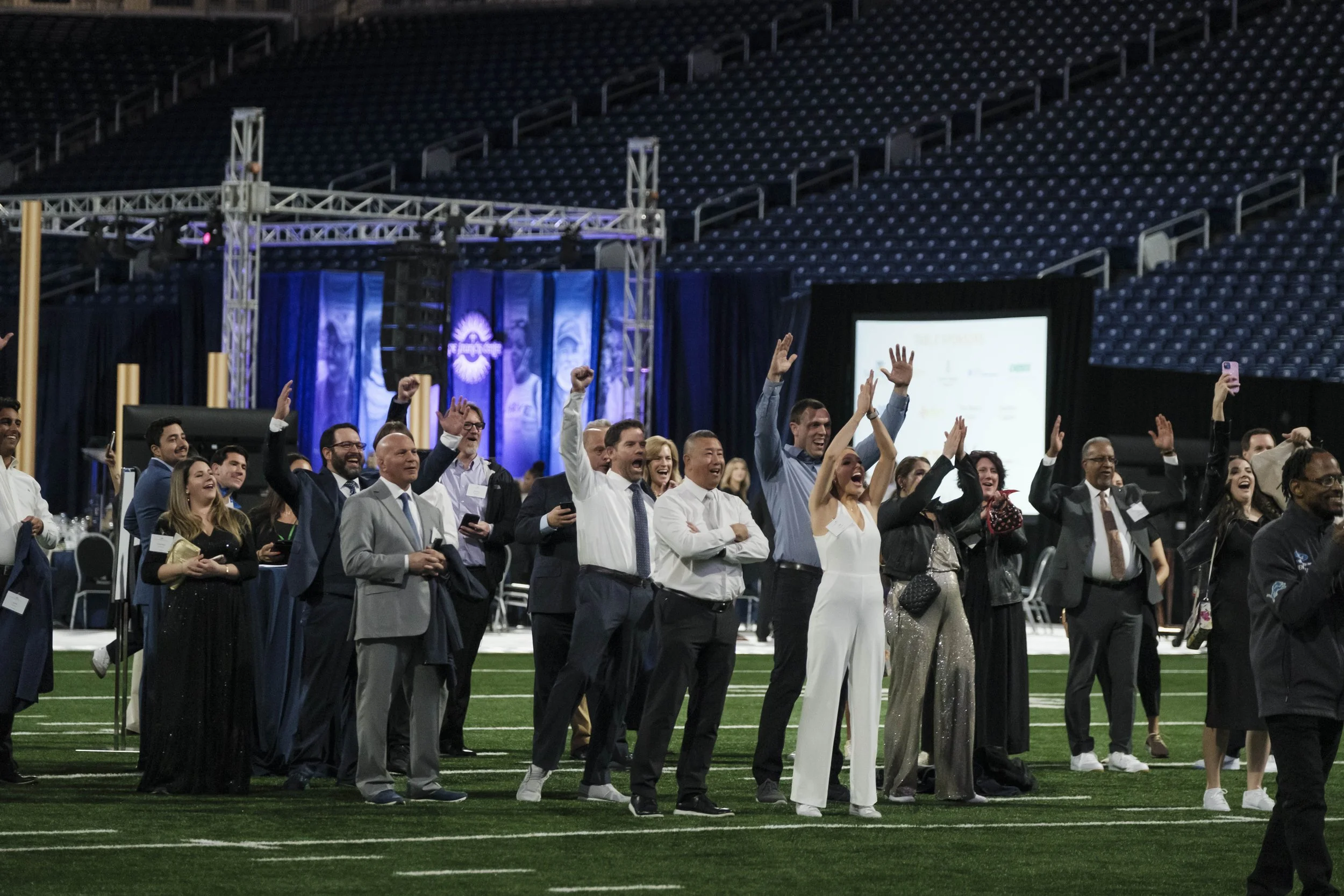 A group of people in suits and gowns cheer on an indoor football field as a group of people stand behind her.