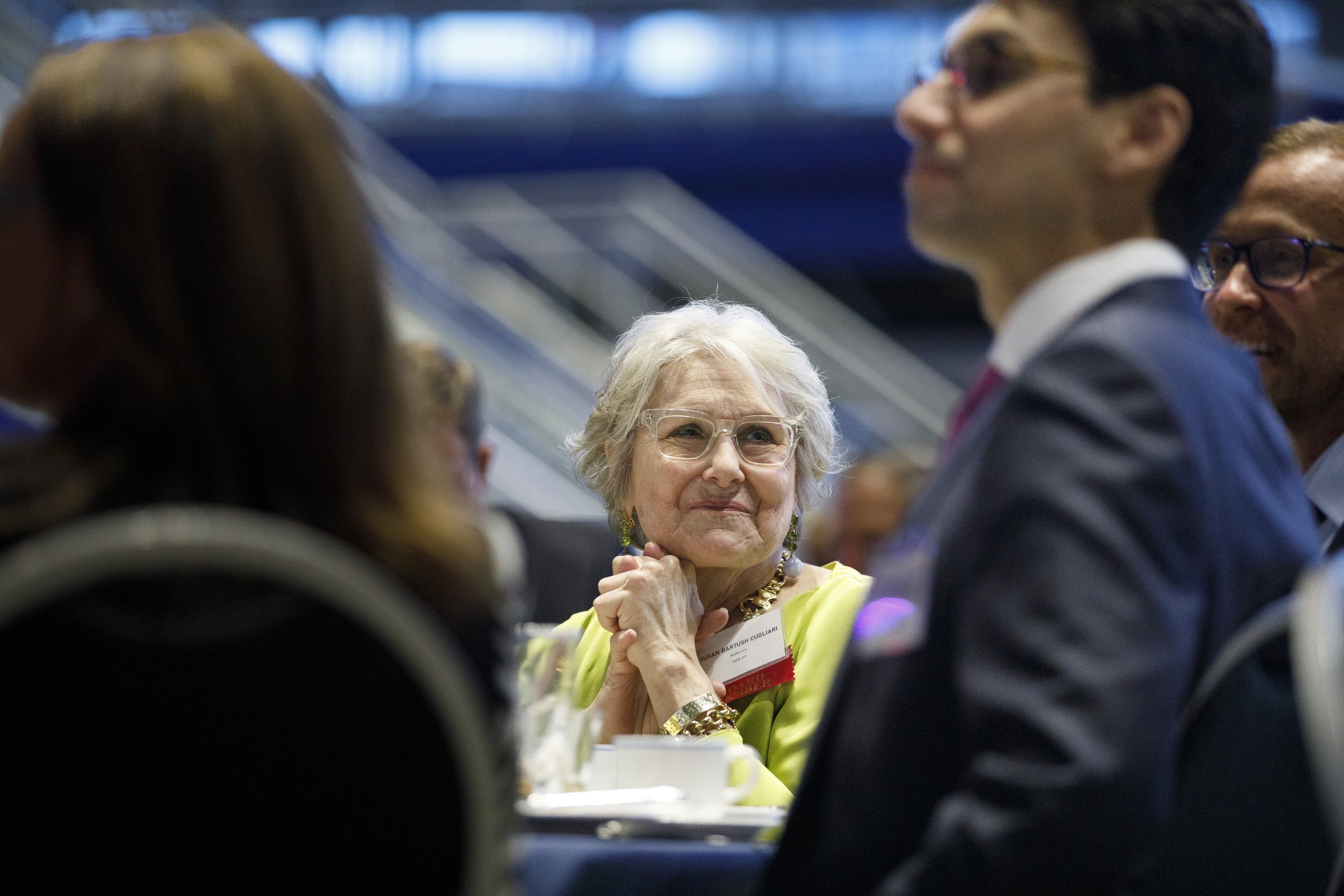 A older woman, with white hair and glasses, sits at a table, holding her hands under her chin while looking off in the distance.