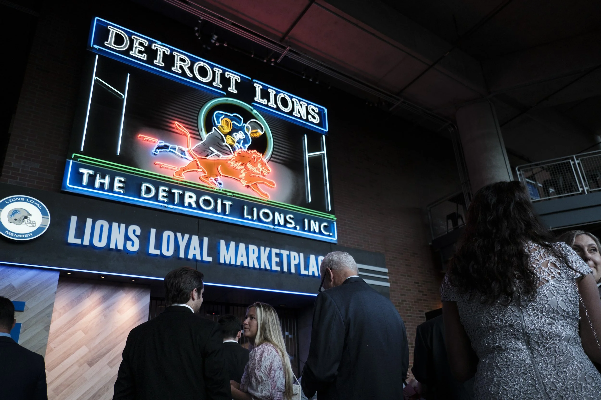A neon sign is illuminated that says "Detroit Lions, The Detroit Lions, Inc" while people stand in the foreground.
