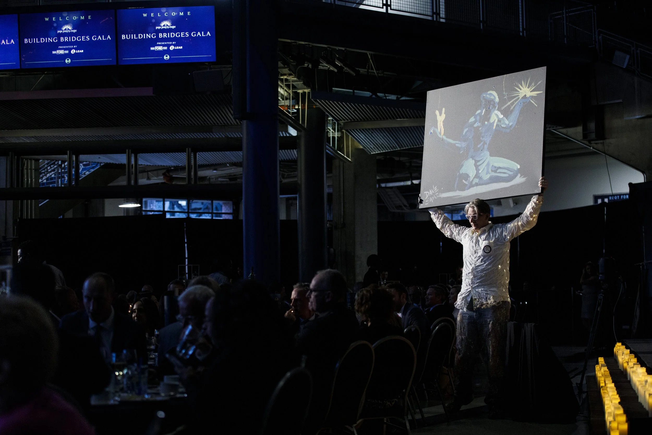 A man stands in a crowd, illuminated by sunlight, while holding up a painting of the Spirit of Detroit.