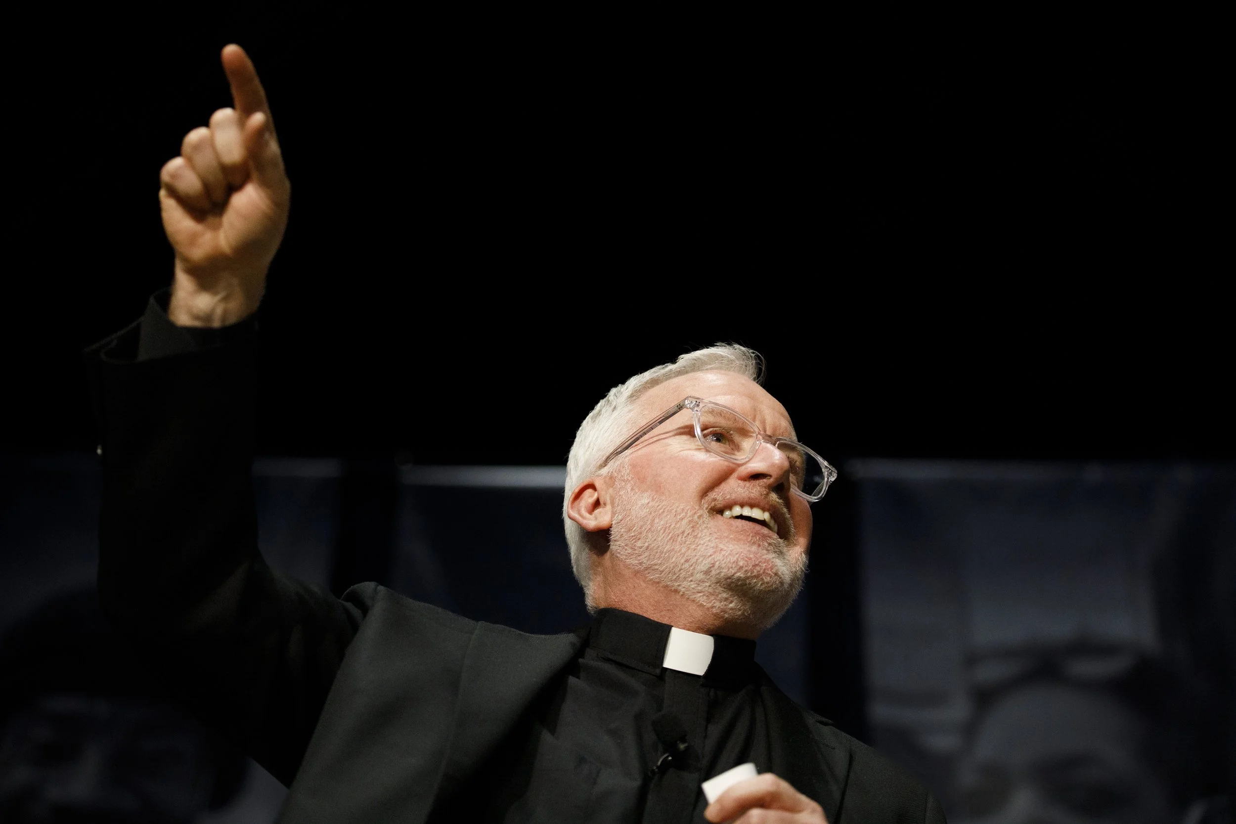 A photo of a man wearing a clerical clothing points up and smiles.