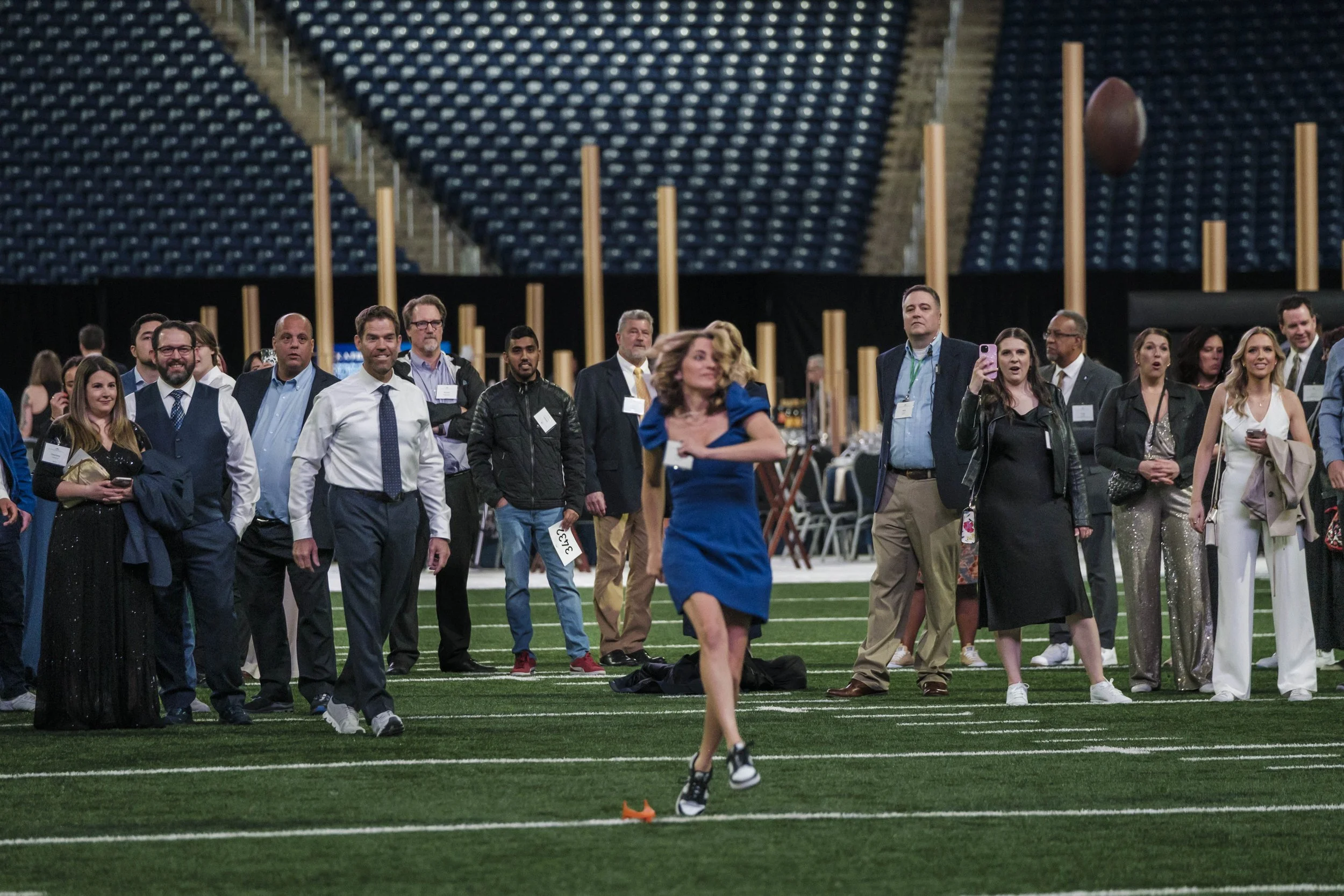A woman in a blue dress kicks a football on an indoor football field as a group of people stand behind her.