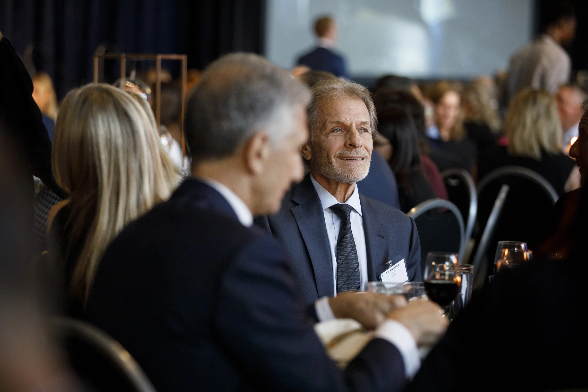 An older man with gray hear and blue eyes smiles while seated at a table.