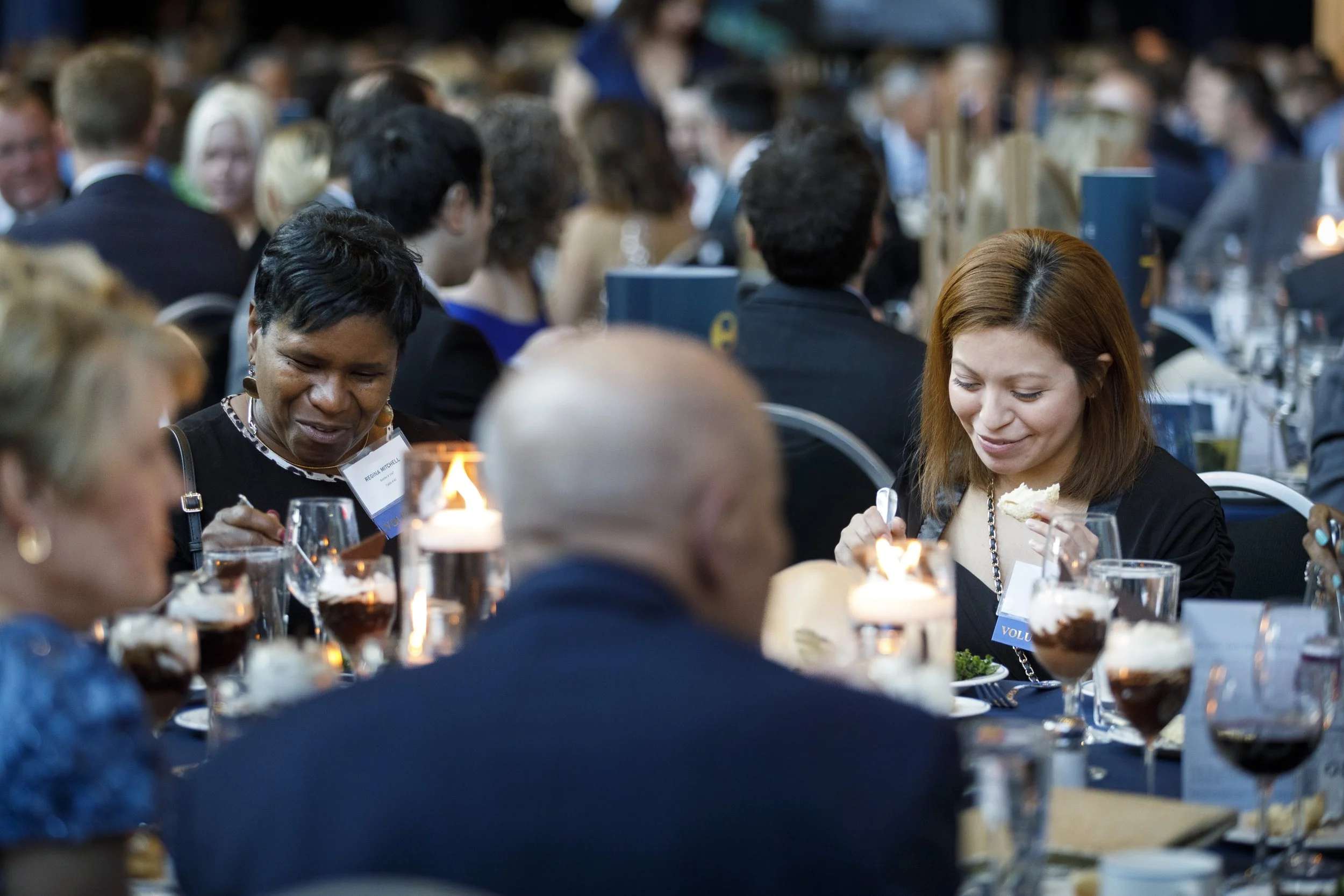 Two women sit at a table and smile while eating food.