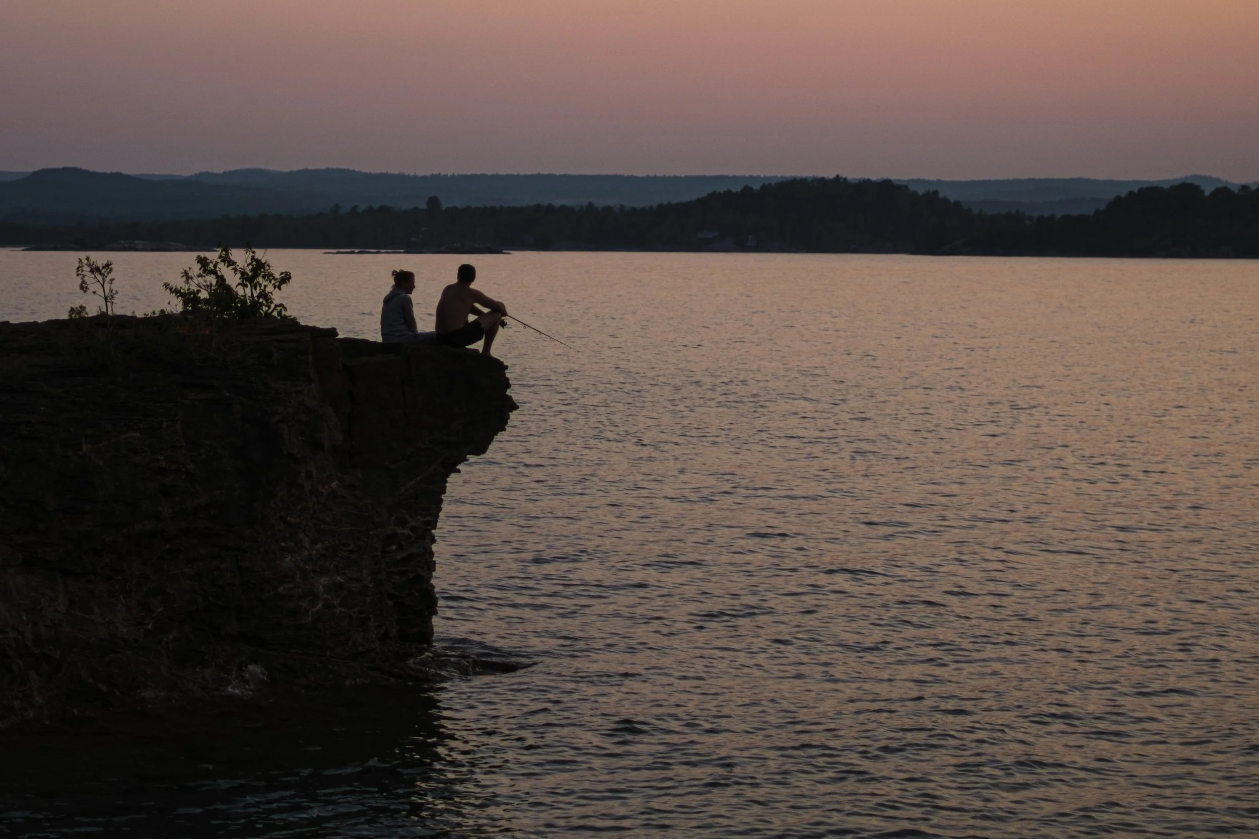 Two people sit silhouetted on a small cliff, one holding a fishing pole,above water with hills and a pink sunset in the background in Marquette, Michigan..