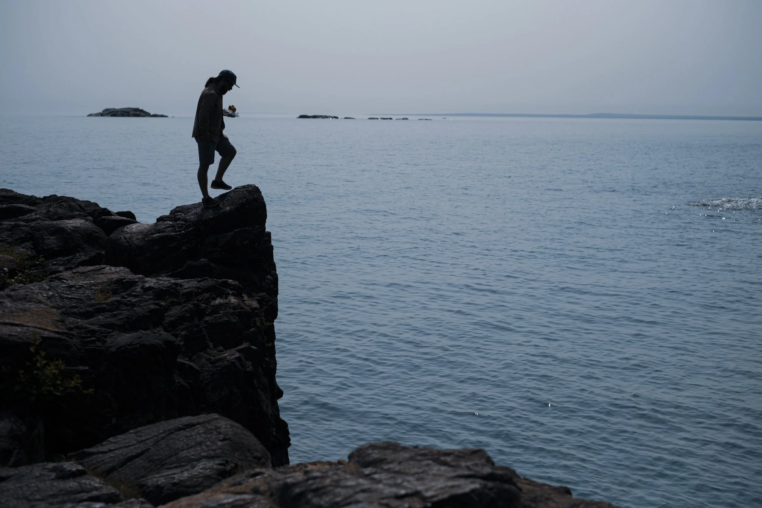 A man stands silhouetted against water while standing on the edge of a cliff.