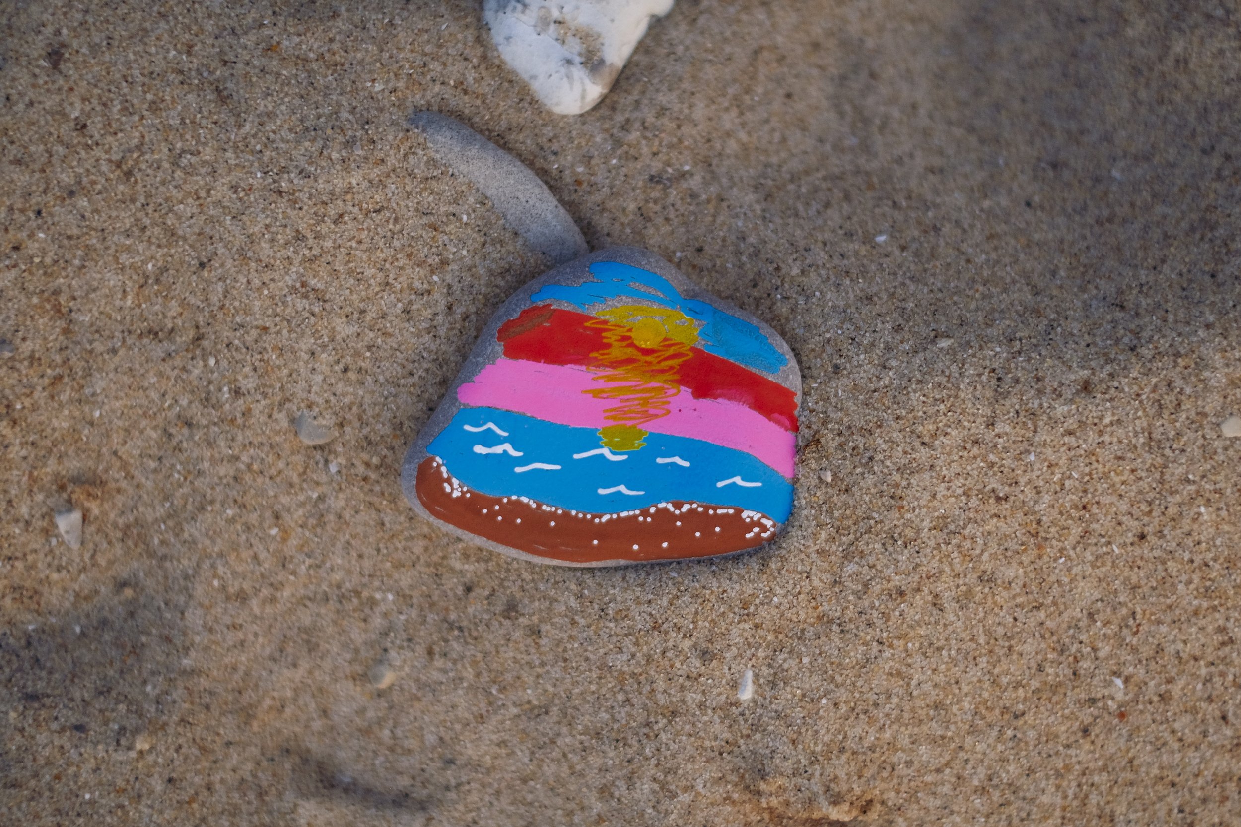 A close up of a colorful, hand painted stone on a beach. The stone has a sunset scene painted on it.
