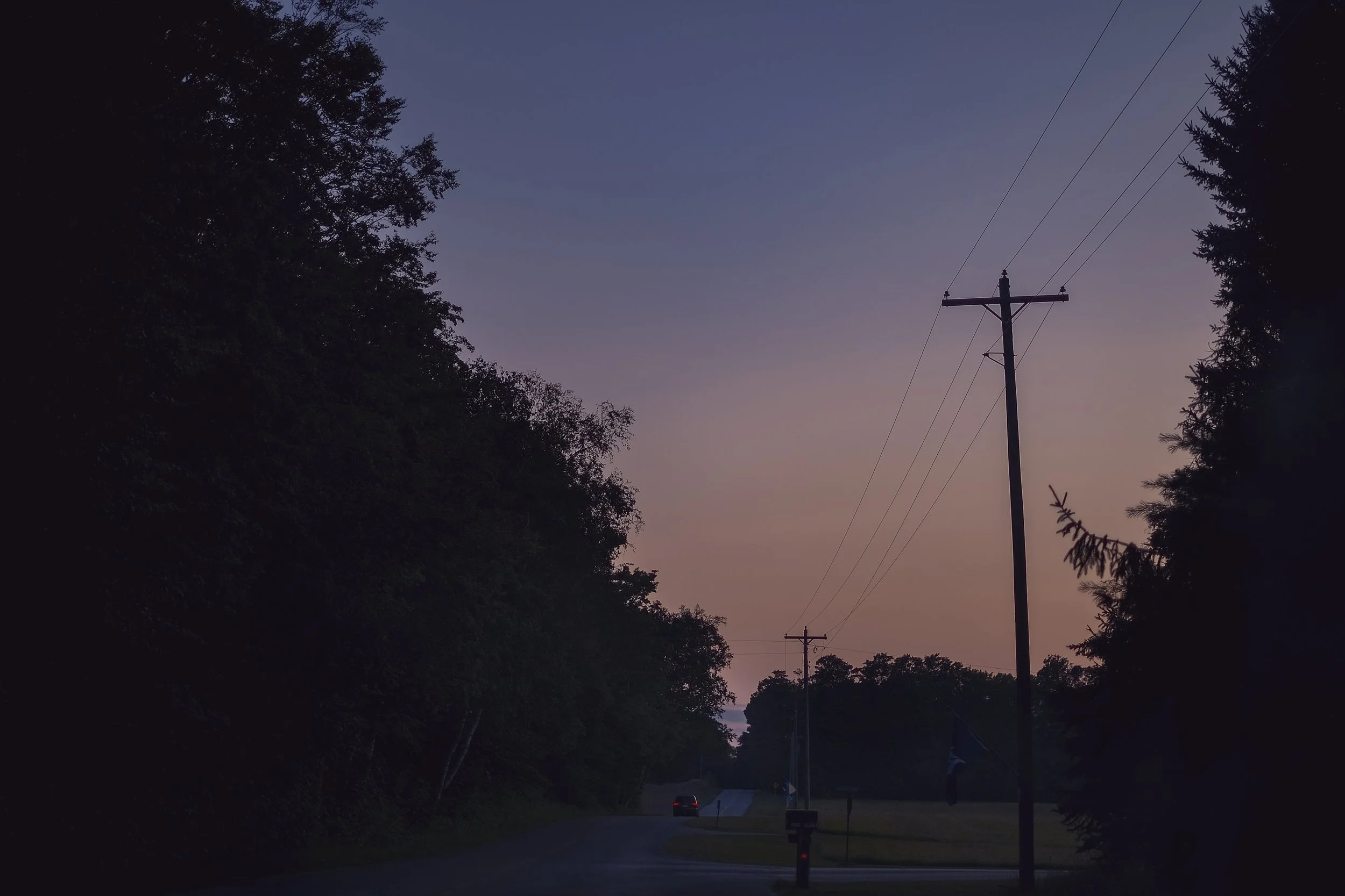 A car with it's brake lights on begins to drives away on a remote road in northern Michigan with power lines silhouetted by a sunset.