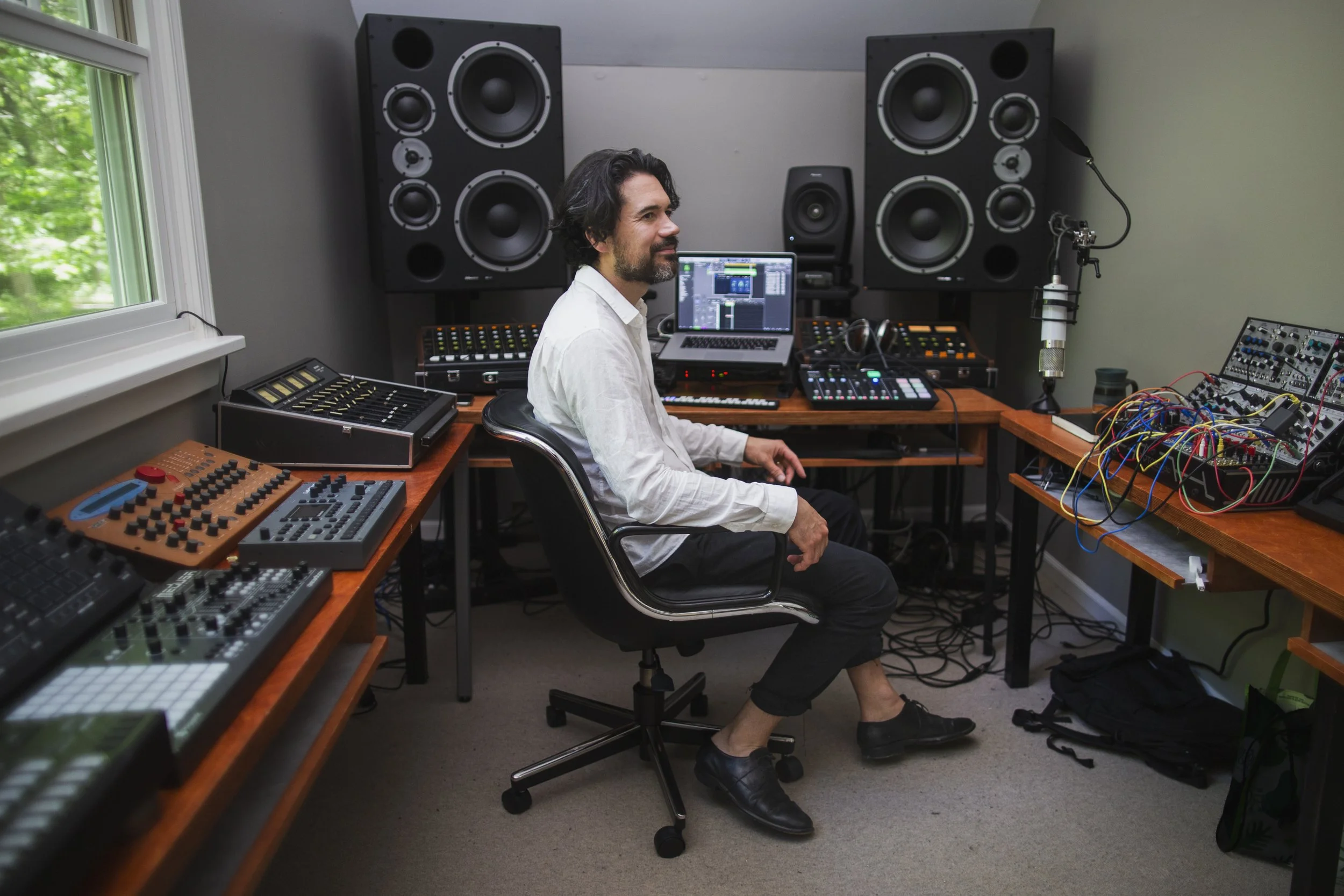 A man sits smiling in a recording studio, surrounded by speakers and other recording equipment.