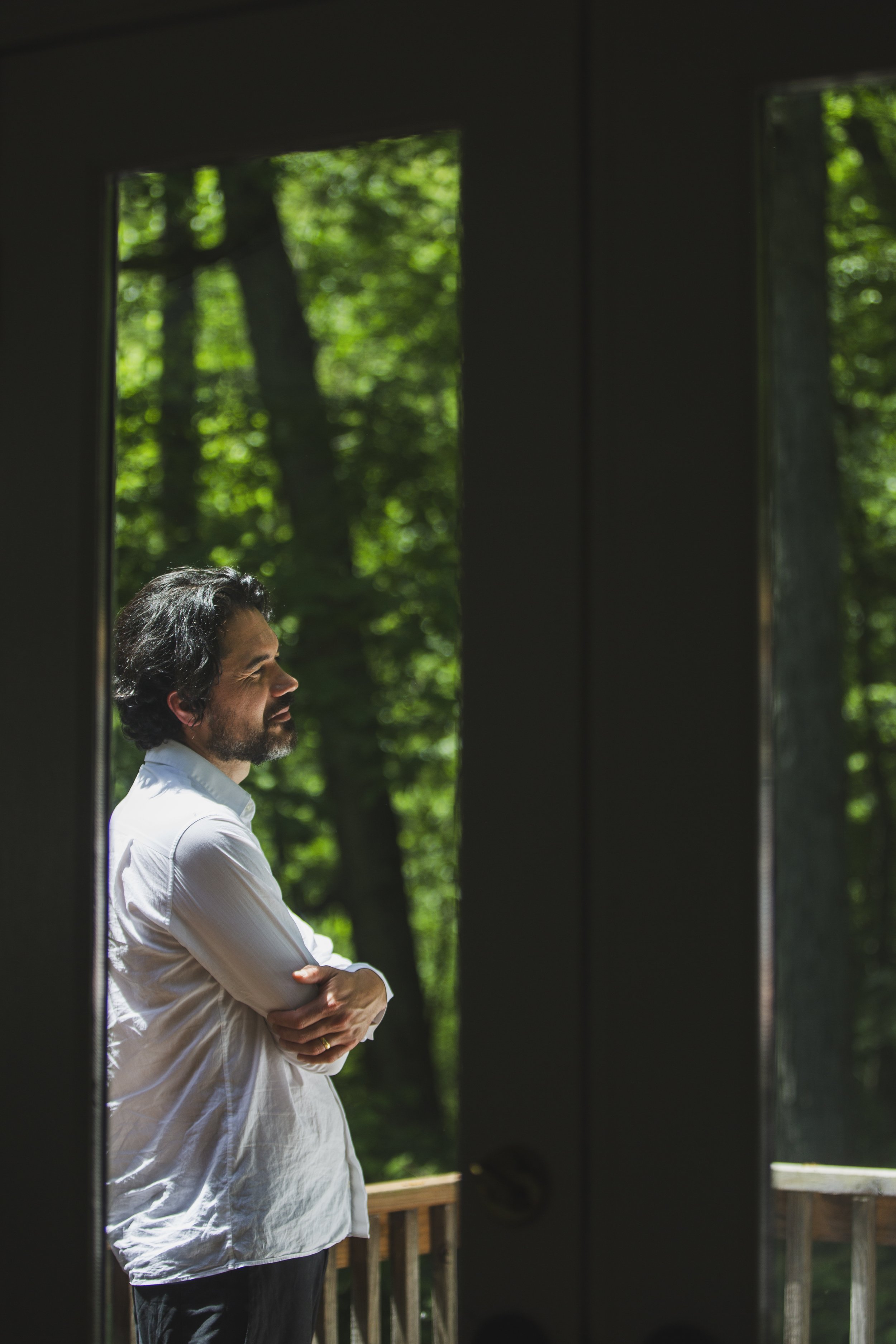 A man standing on a balcony with his arms crossed, outdoors, surrounded by trees.