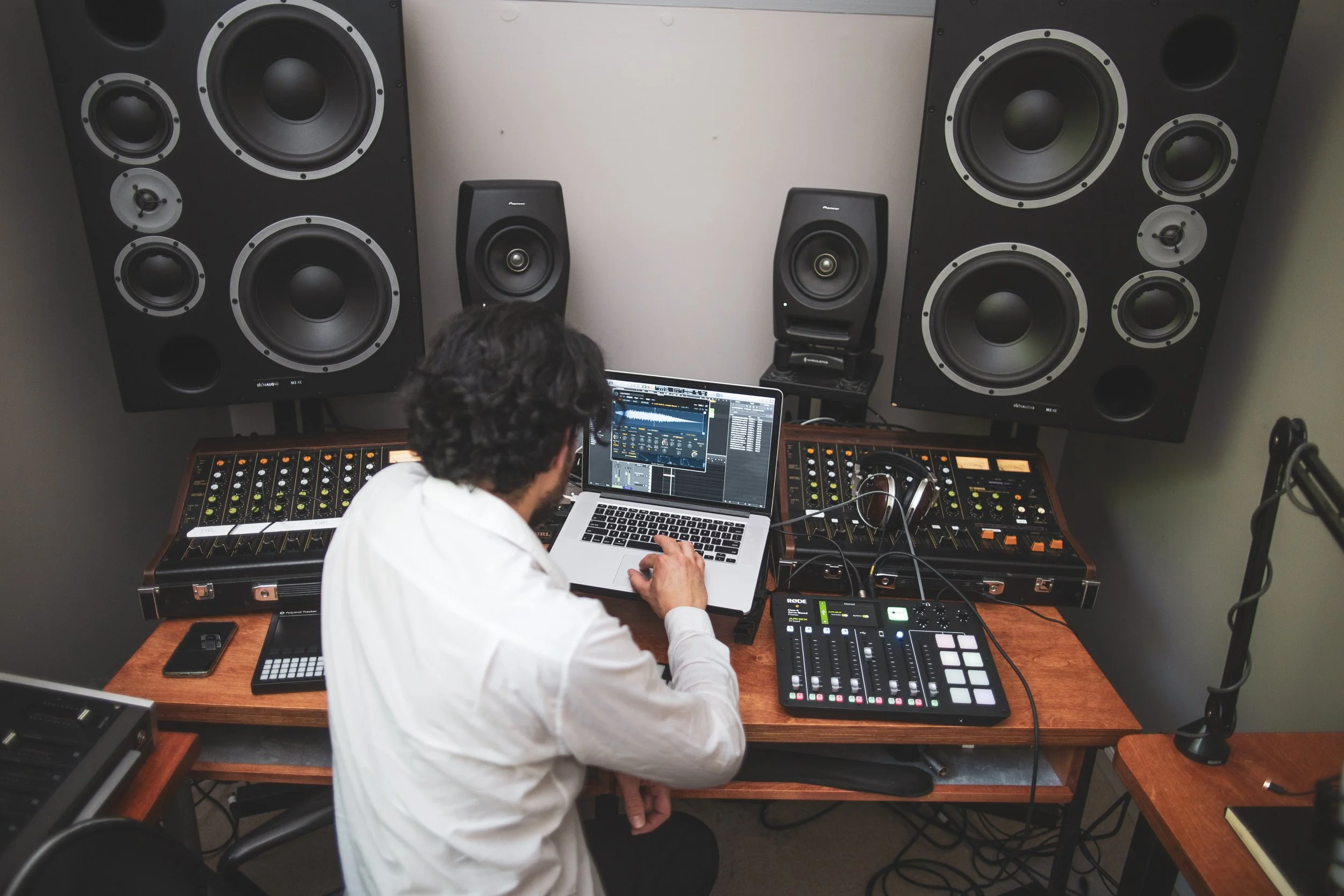 A man sits with this hands on a laptop in a recording studio, surrounded by speakers and other recording equipment.