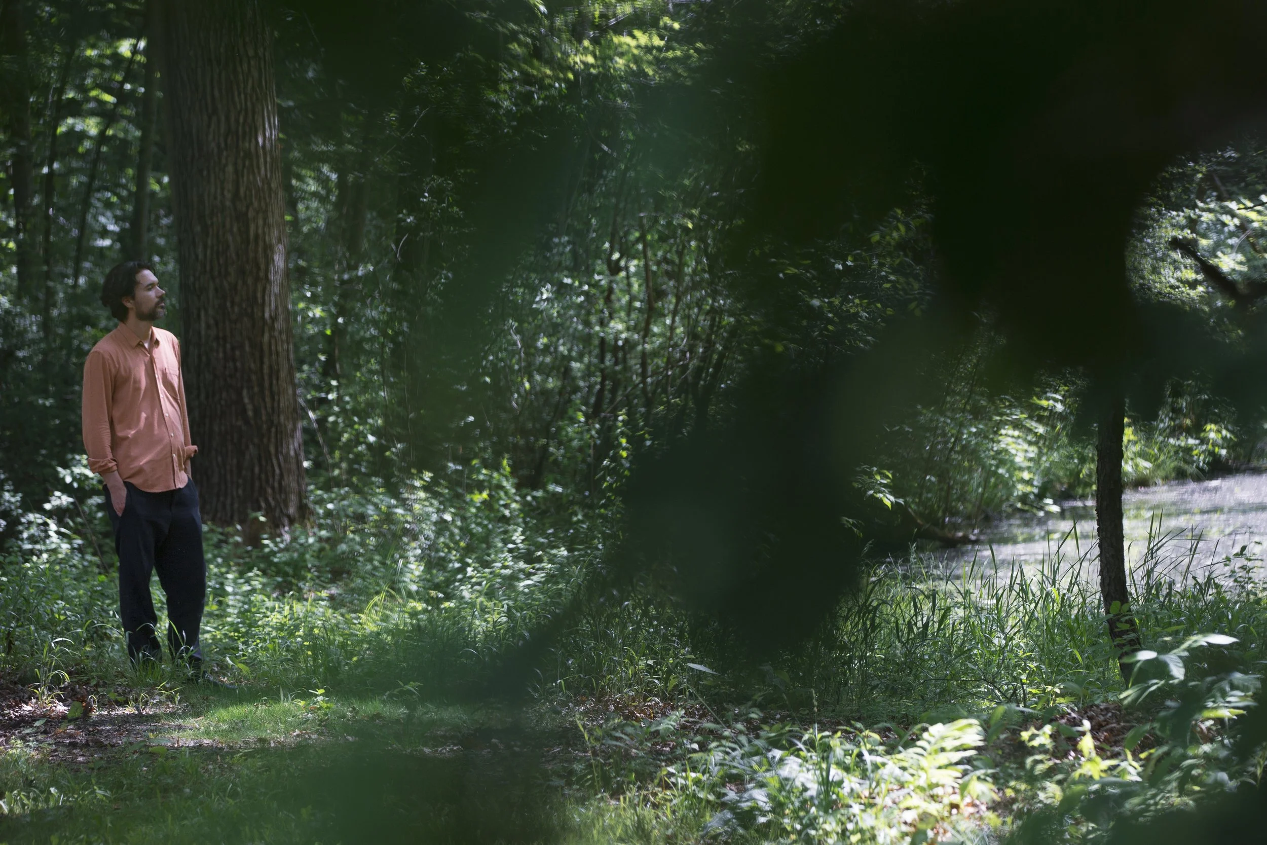 A man stands near the edge of a pond, surrounded by trees, framed between blurry leaves.