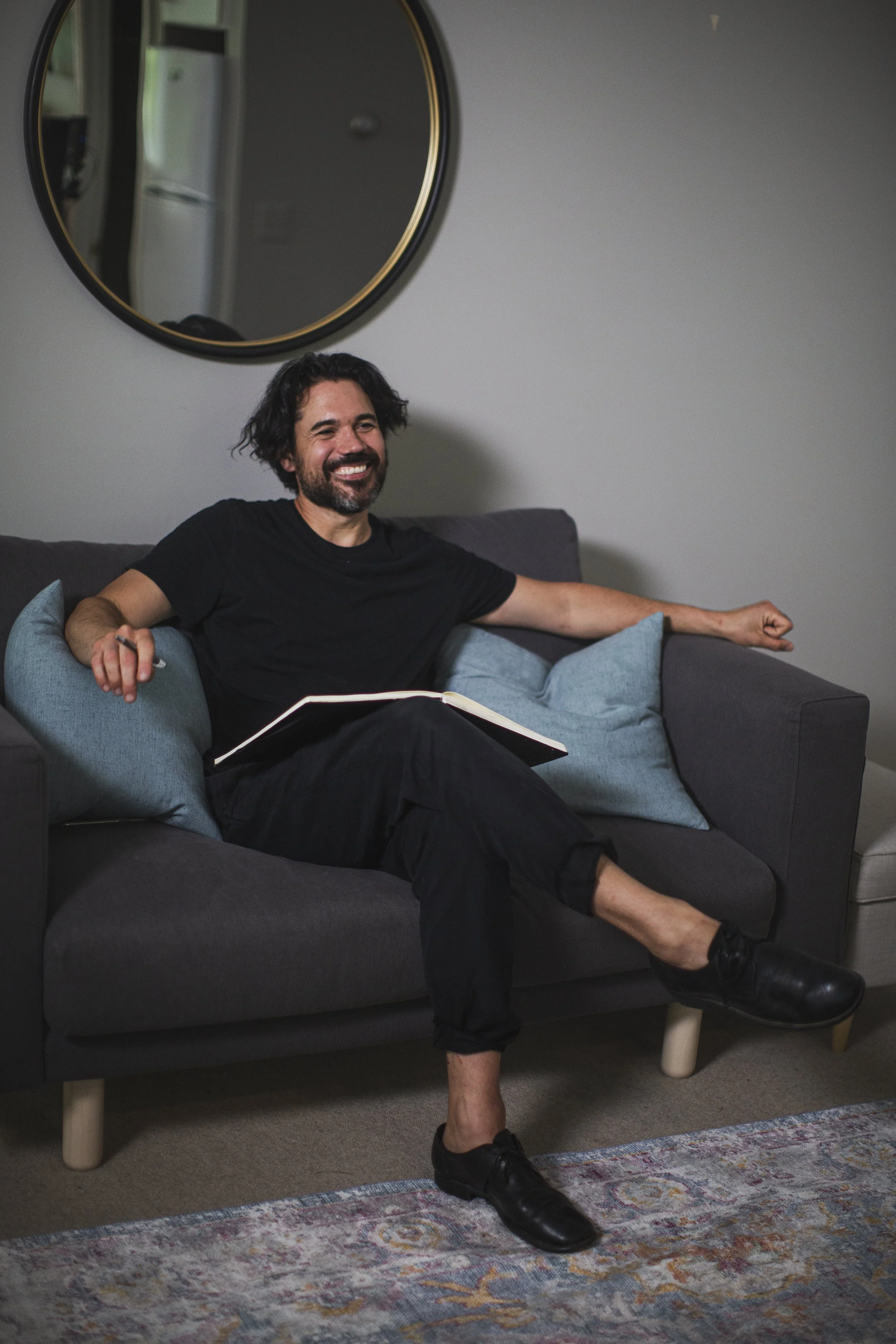 A man smiles while sitting on a chair with his legs crossed and holding a pen and a notebook in his lap.