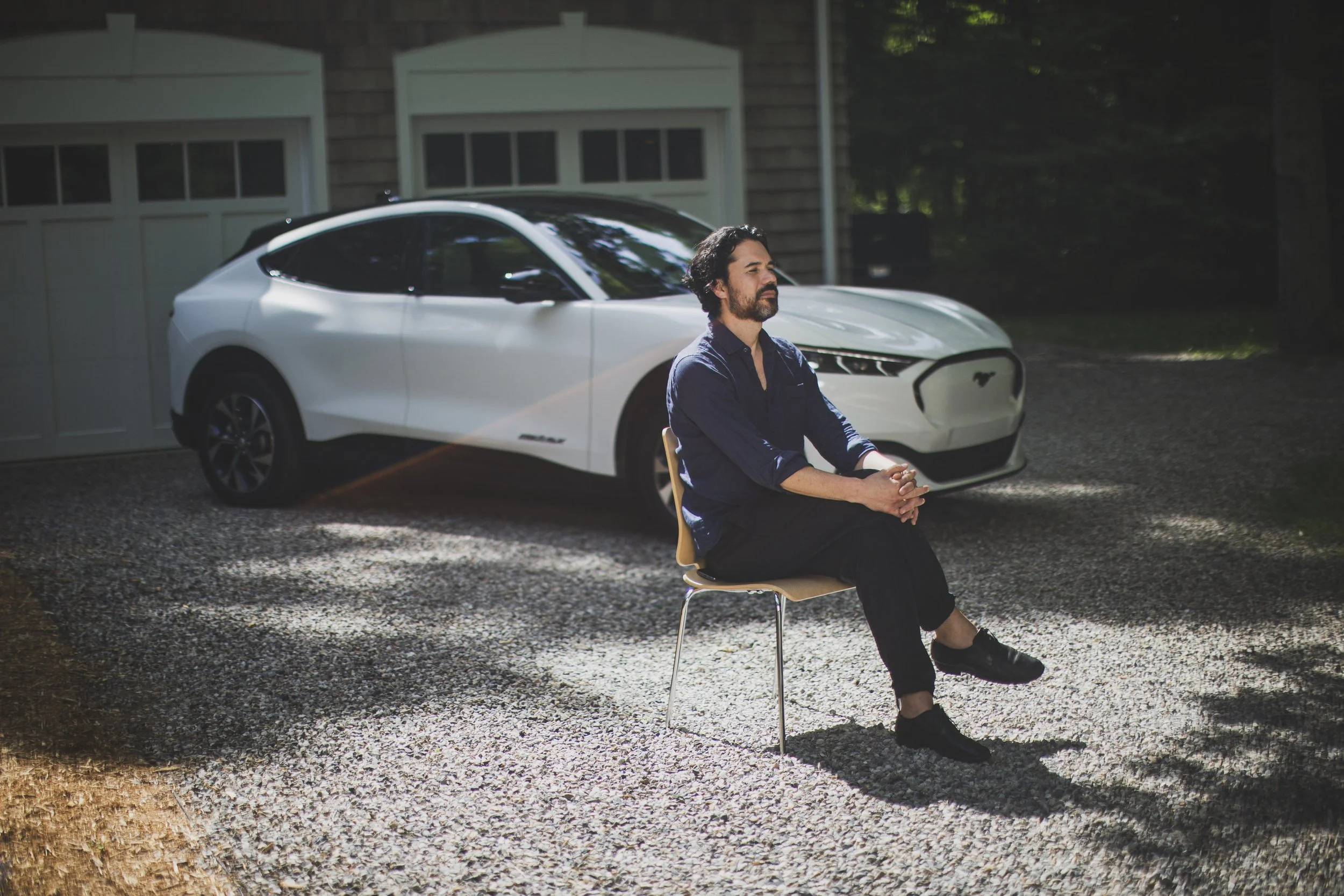 A man sits outdoors in a chair, in the middle of a driveway, with a white car in the background.