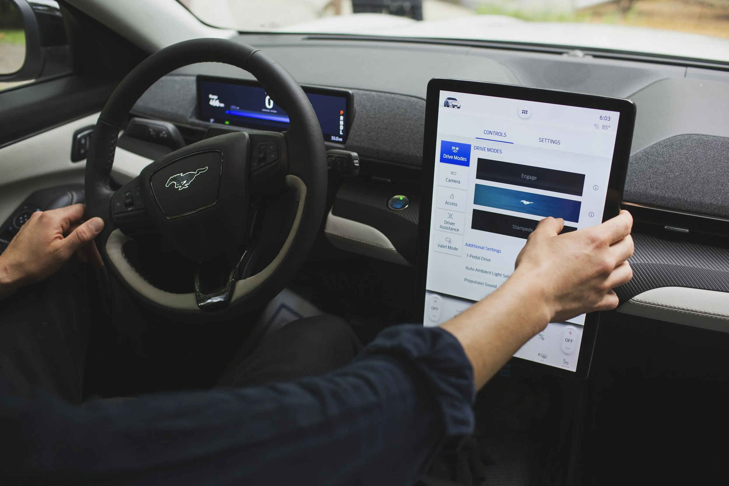 A hand reaches out to use a touch screen in a car while also holding a steering wheel with a mustang emblem in the center.