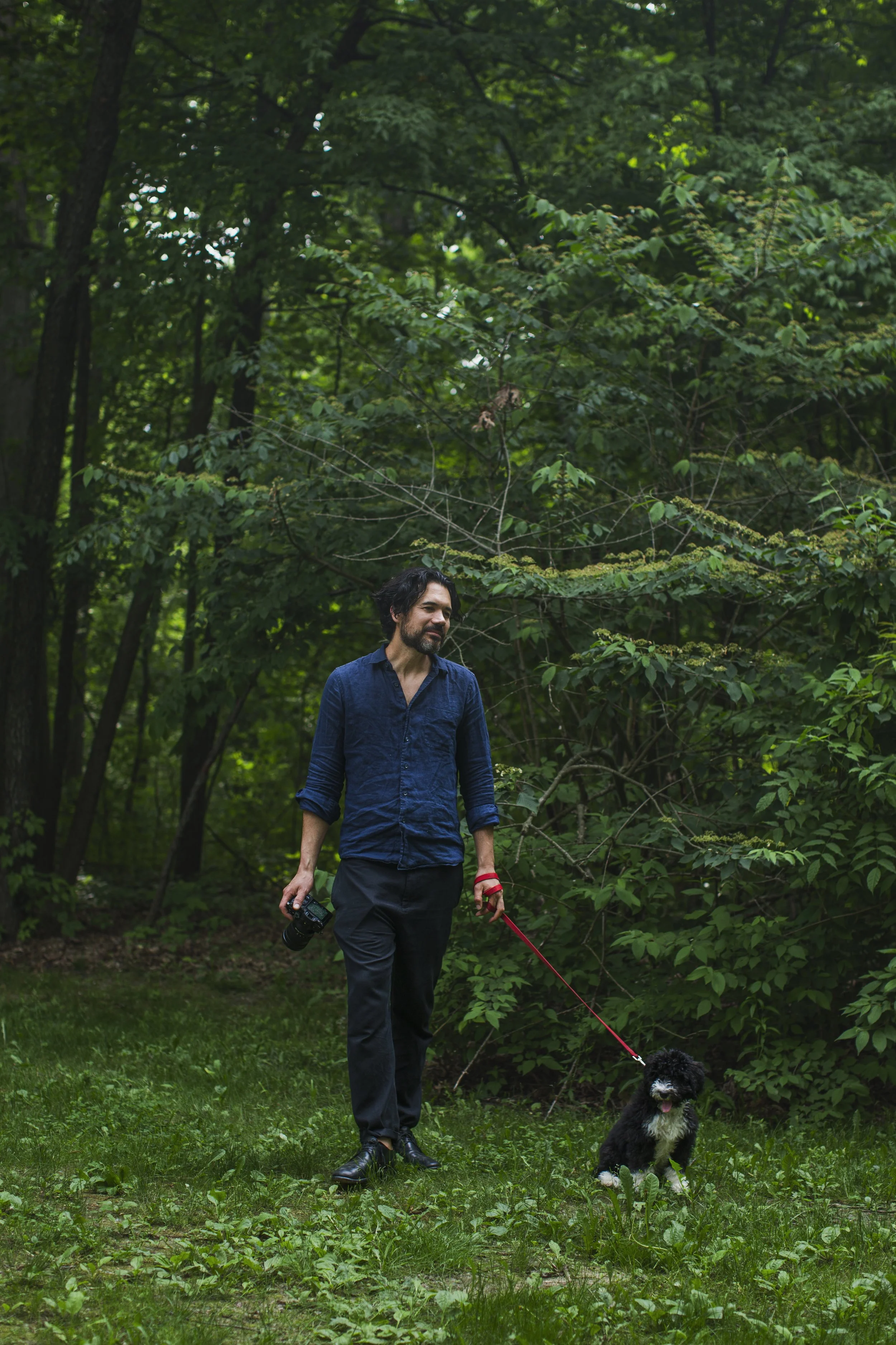 A man stands, holding a camera in one hand and red dog leash in another, in the middle of a green forest.