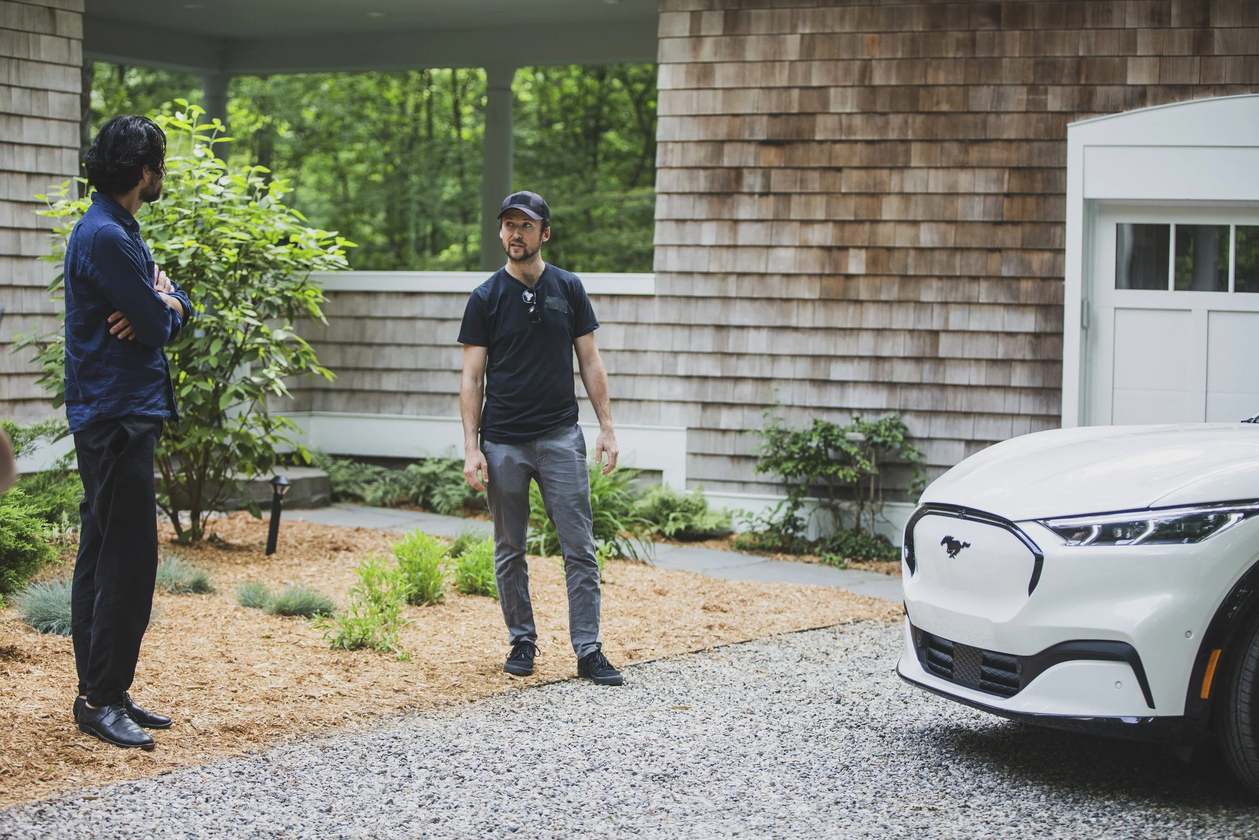 Two men stand in a driveway with a white car, talking to one another.