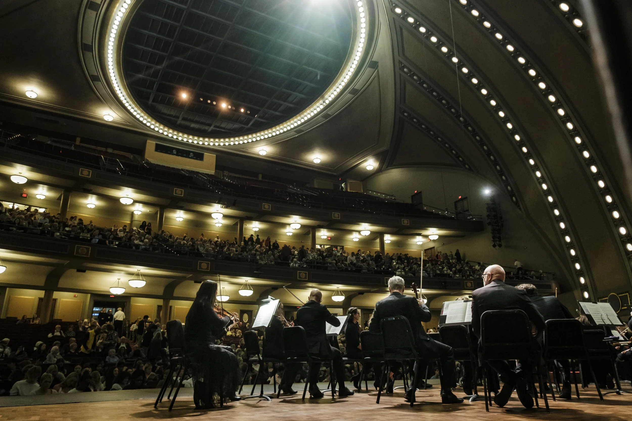 The Ann Arbor Symphony Orchestra on stage with their instruments, facing the audience with bright stage lighting.