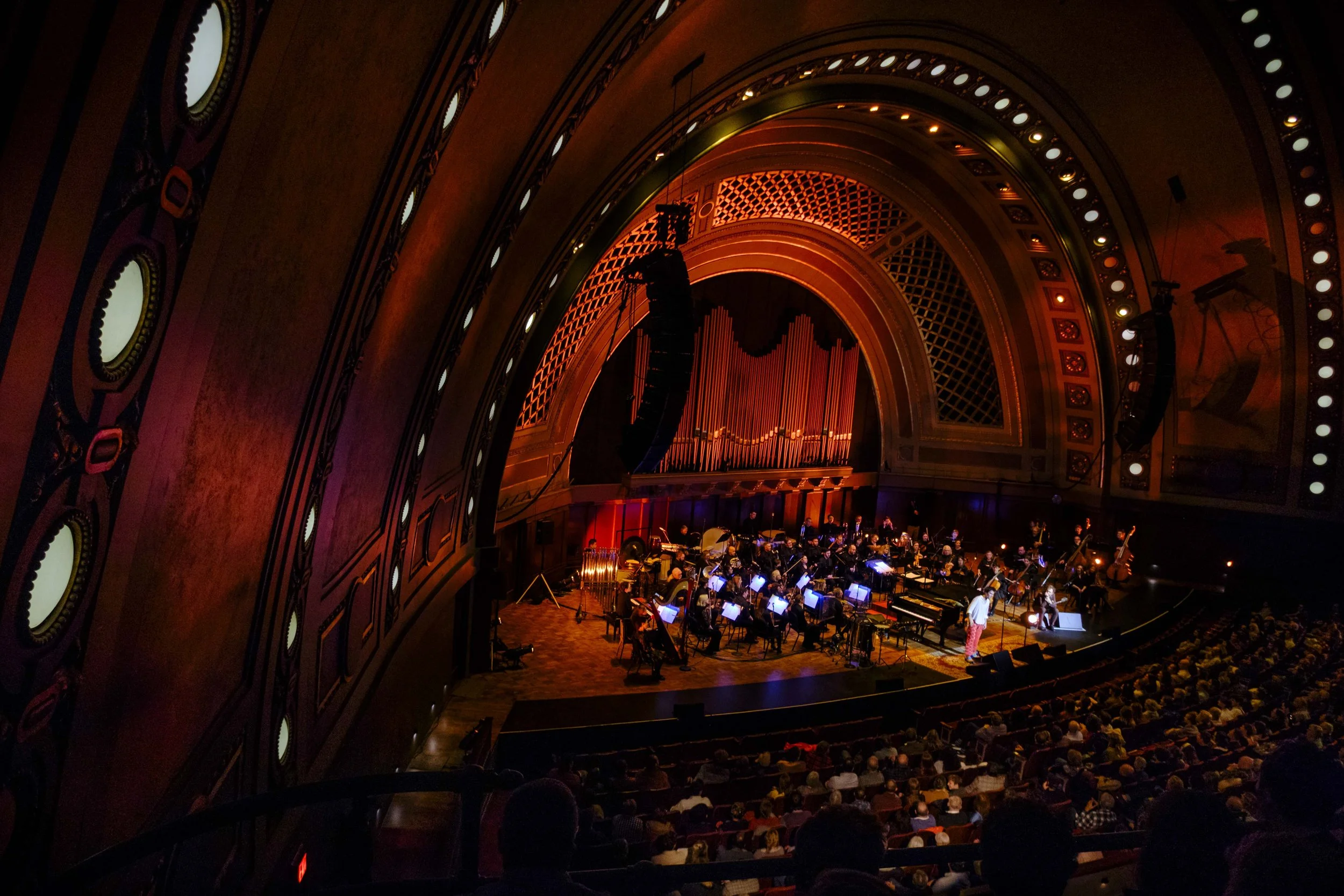 Jacob Collier performing a concert with the Ann Arbor Symphony Orchestra on stage with orange concert lighting.