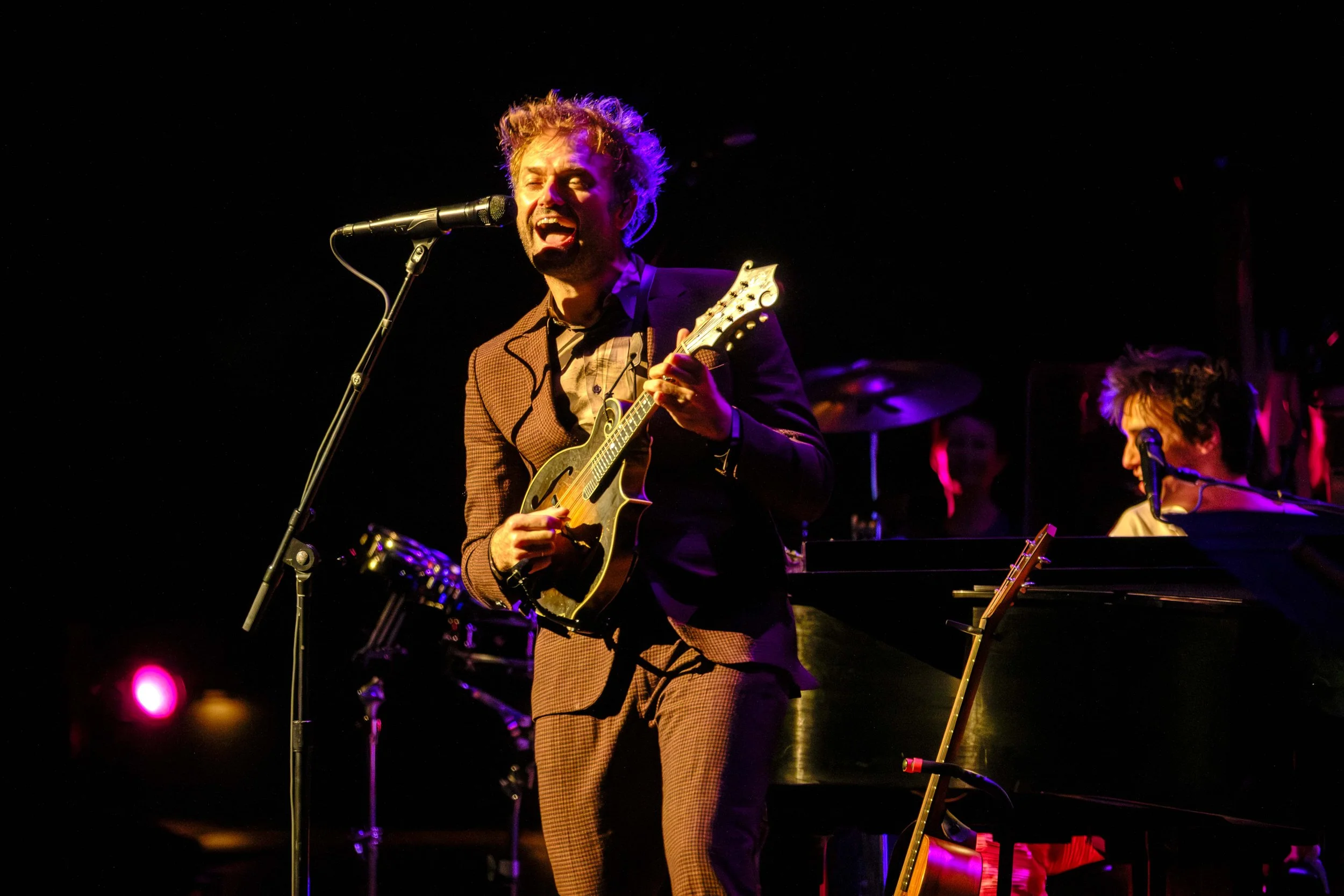 Chris Thile singing into a microphone during a concert with the Ann Arbor Symphony Orchestra with orange and purple stage lighting.
