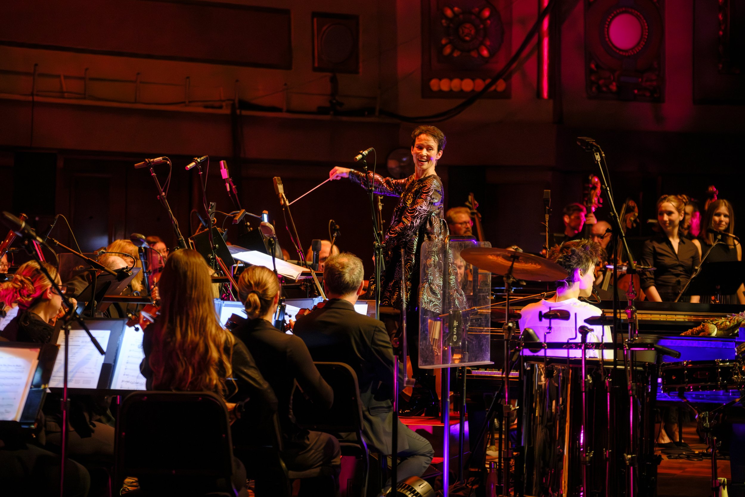 Suzier Collier conducting the Ann Arbor Symphony Orchestra on stage with purple and orange stage lighting.