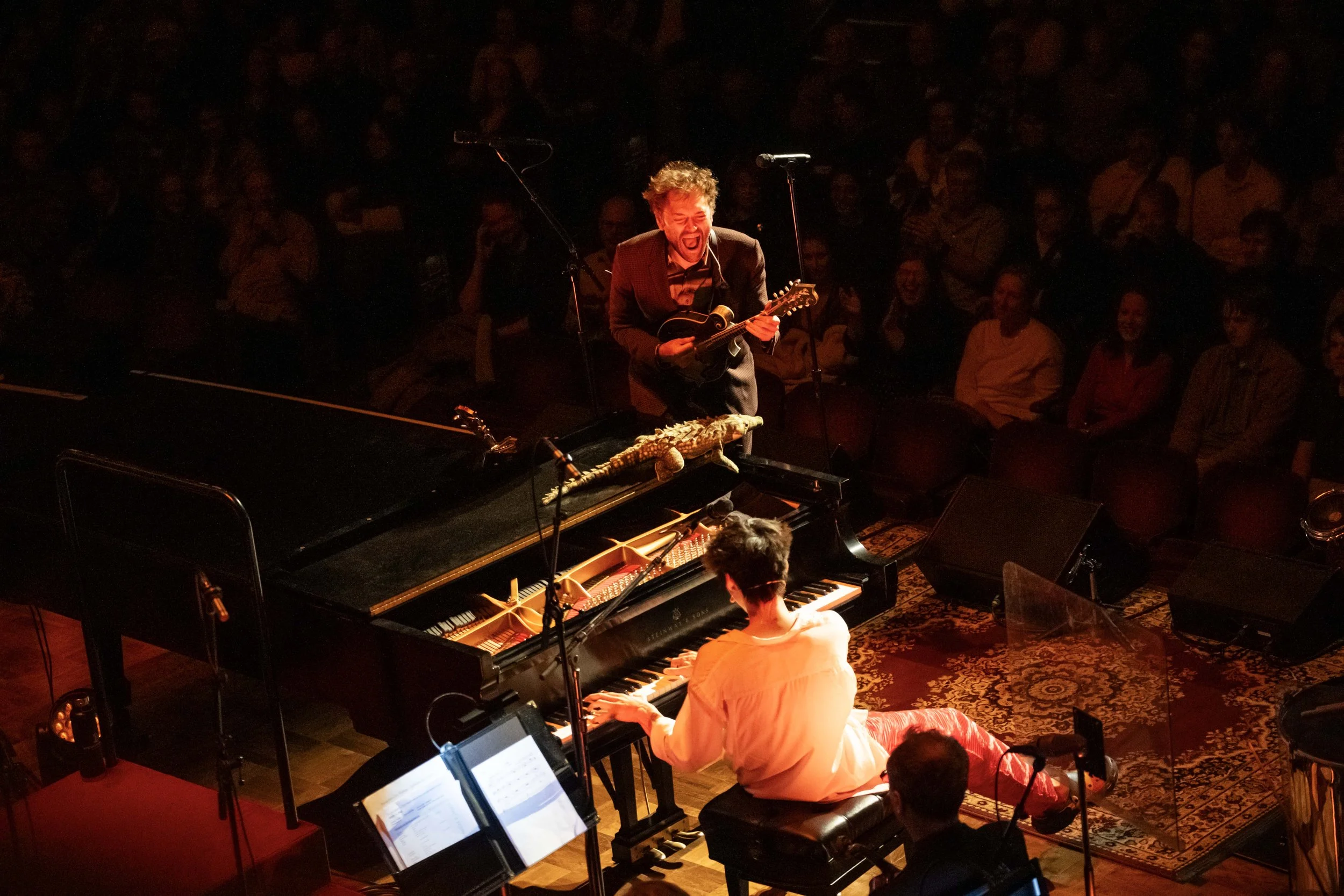Chris Thile sings during a concert with the Ann Arbor Symphony Orchestra with red and orange stage lighting.