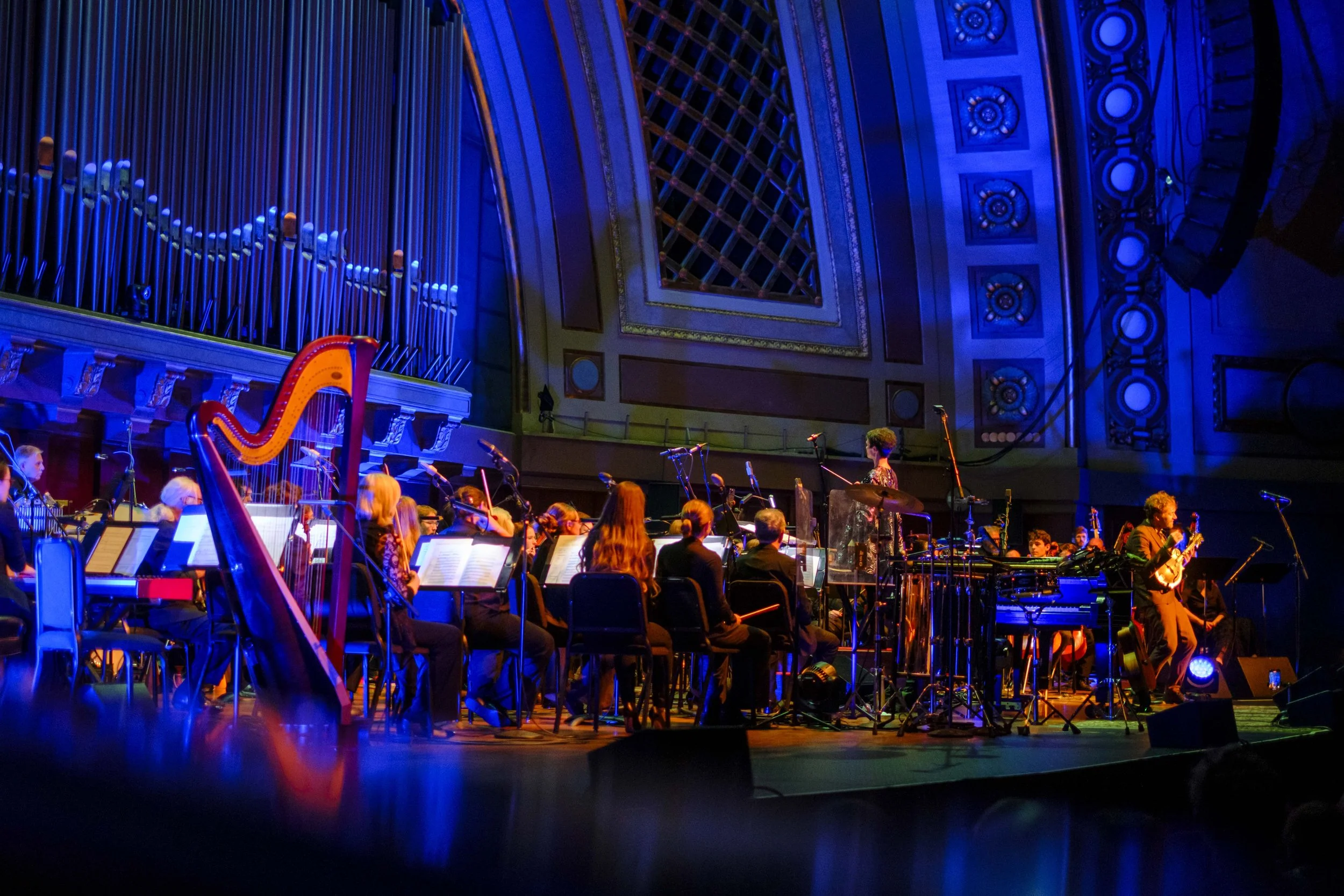 Chris Thile performing on stage during a concert with the Ann Arbor Symphony Orchestra with multi-colored stage lighting.