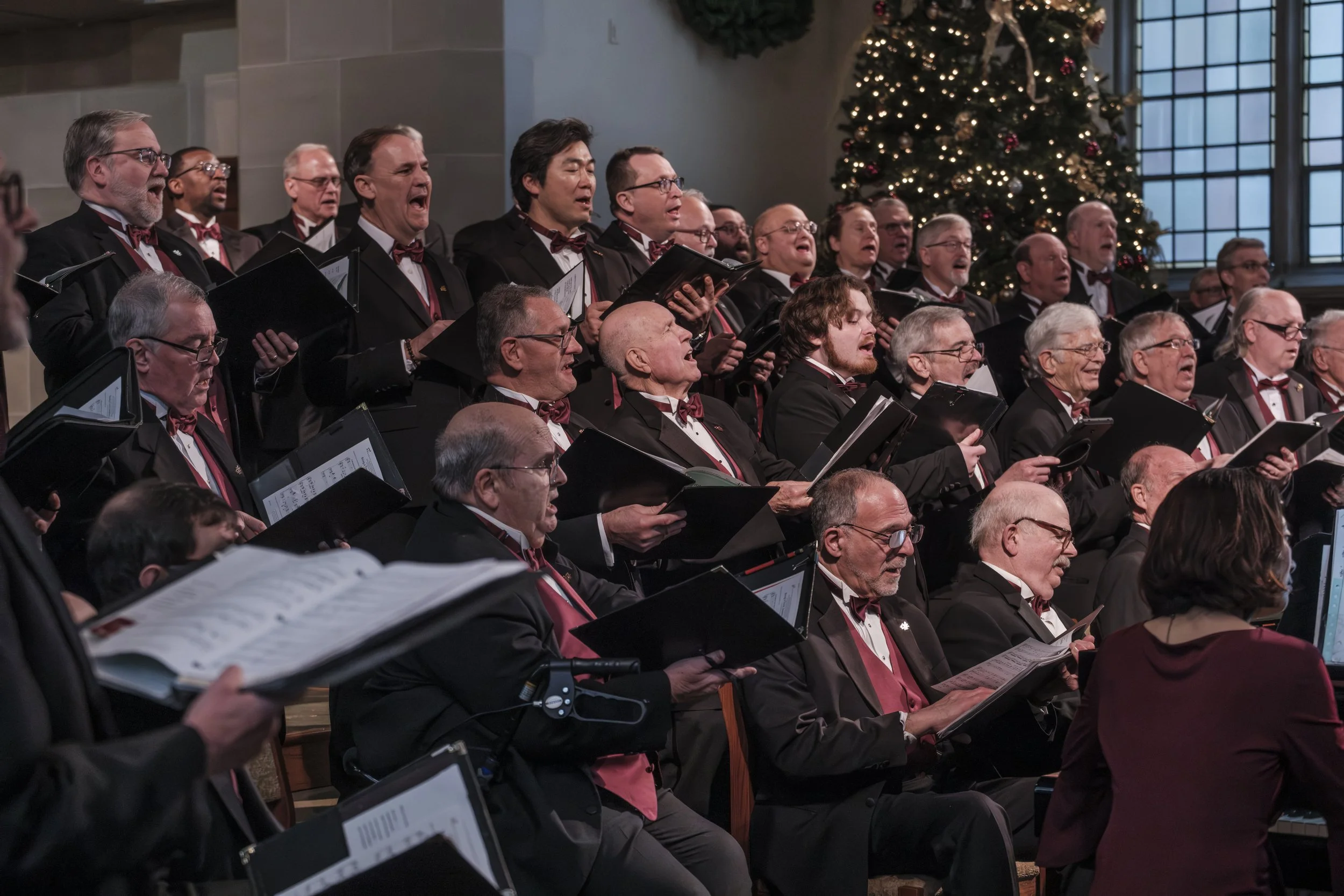 A group of men singing while holding binders with a christmas tree in the background.