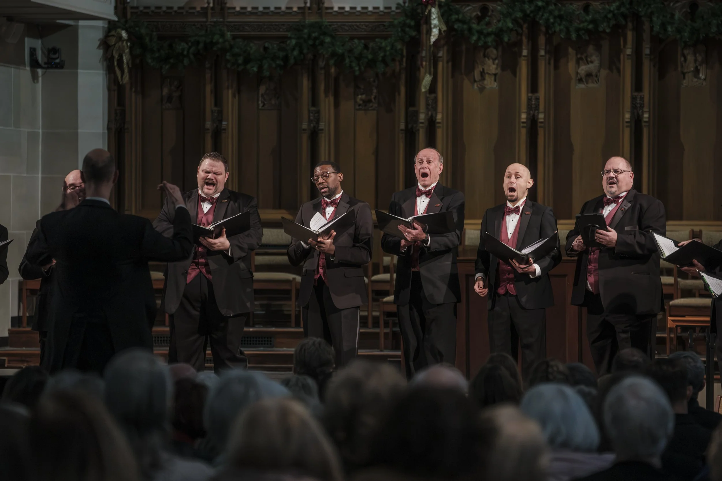 Six men stand singing while a conductor, who has his back to the audience, directs them.