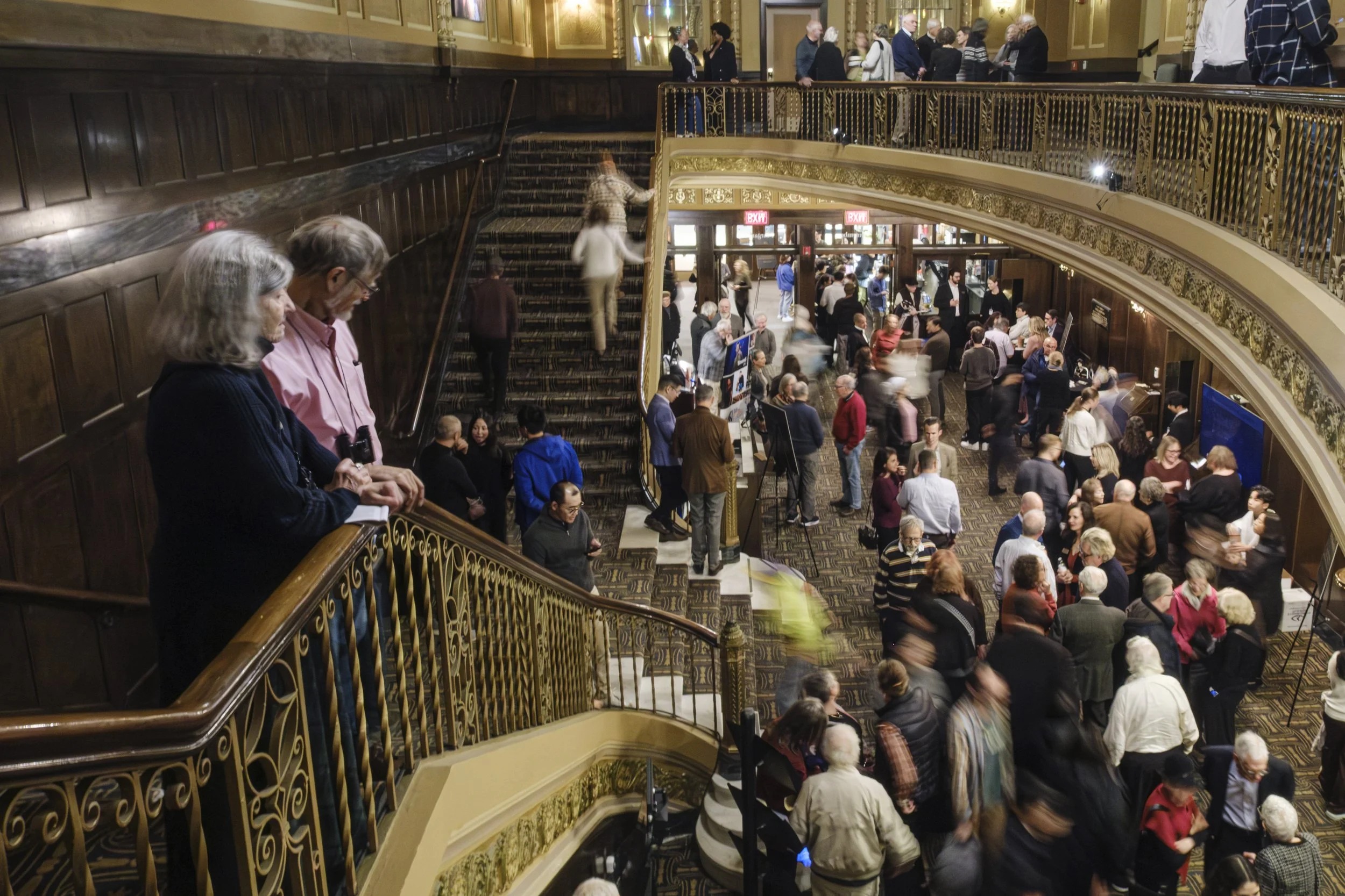 A couple standing and watching people from the balcony as blurred people move around in a lobby.