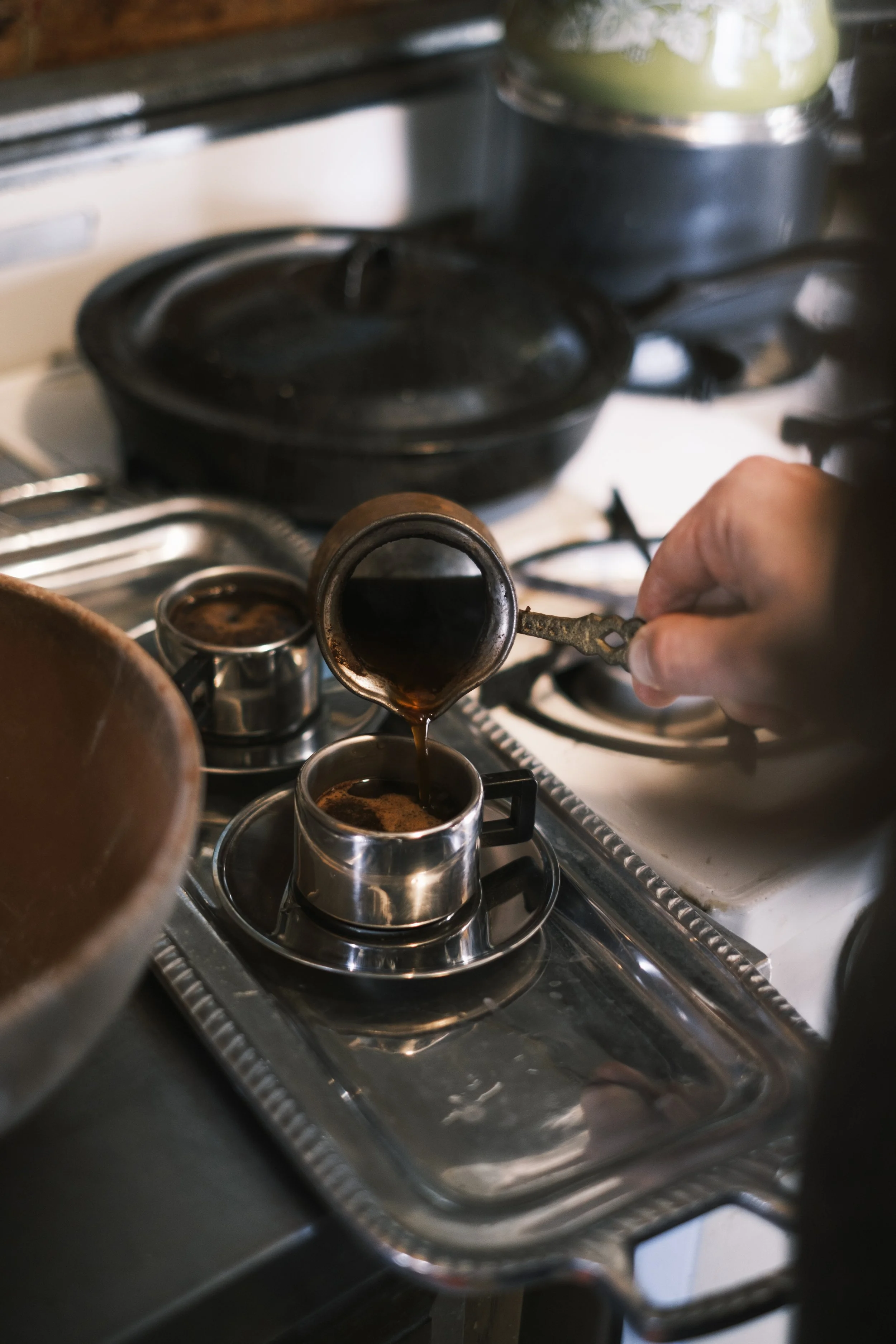 A hand pours coffee from a carafe into a cup with a stove in the background.