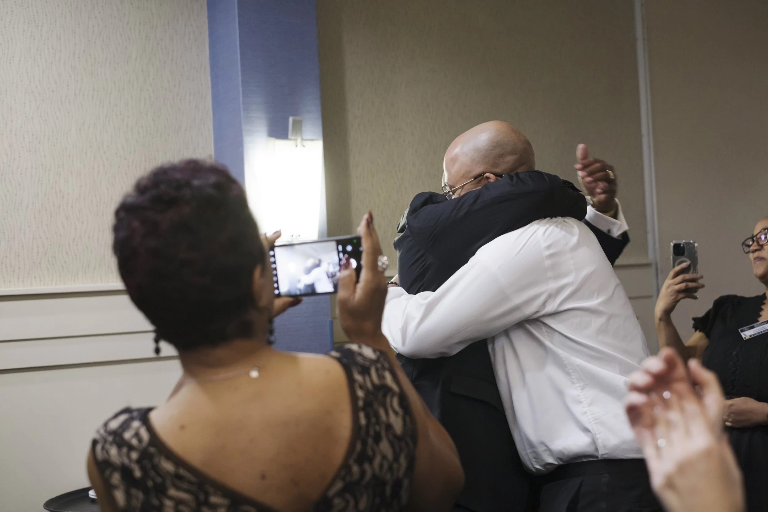 Two people hug while a person in the foreground takes a photo on her cellphone.