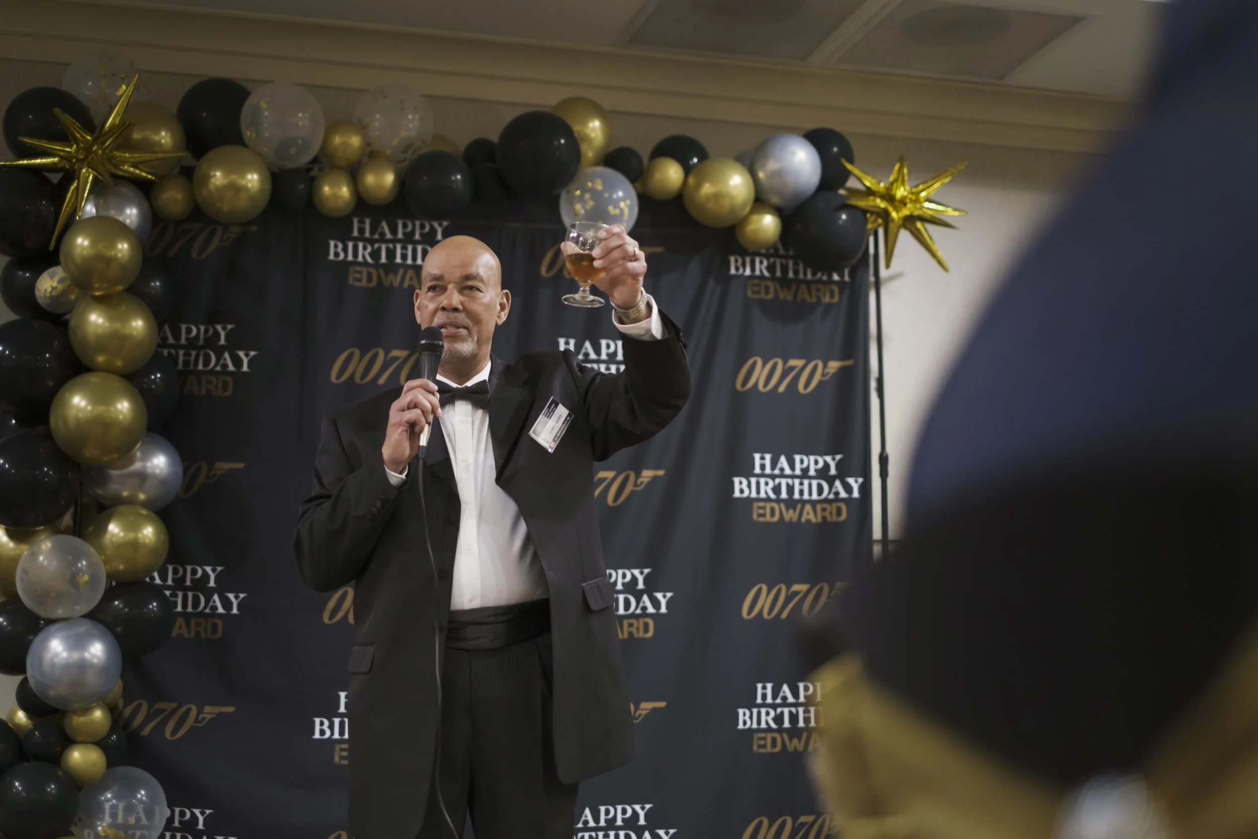 A man wearing a suit hold up a glass while holding a microphone.