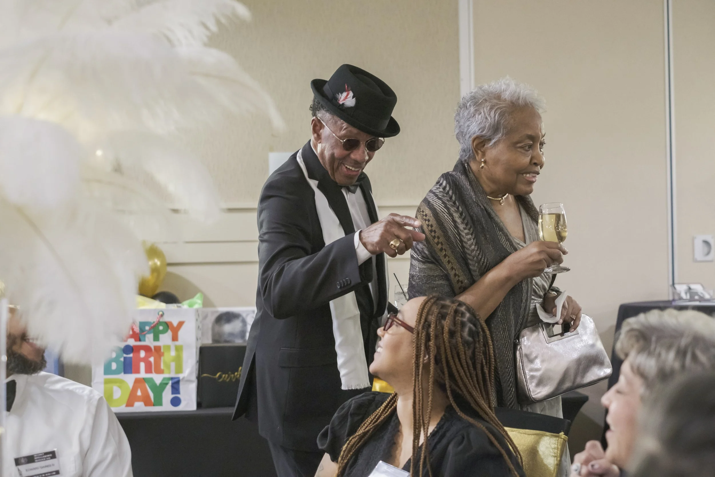 Two people, one in a dress holding a wine glass and another wearing a suit with a hat and sunglasses points to a person sitting at a table.