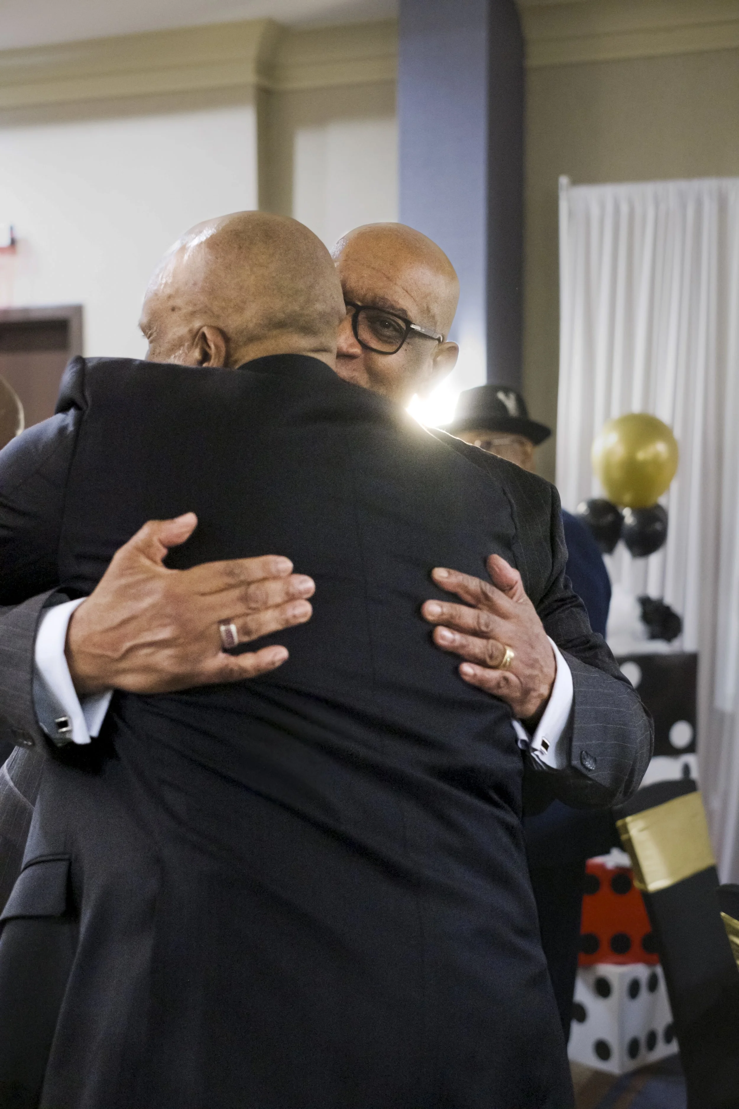 A man wearing glasses hugs another man in a suit with balloons in the background.