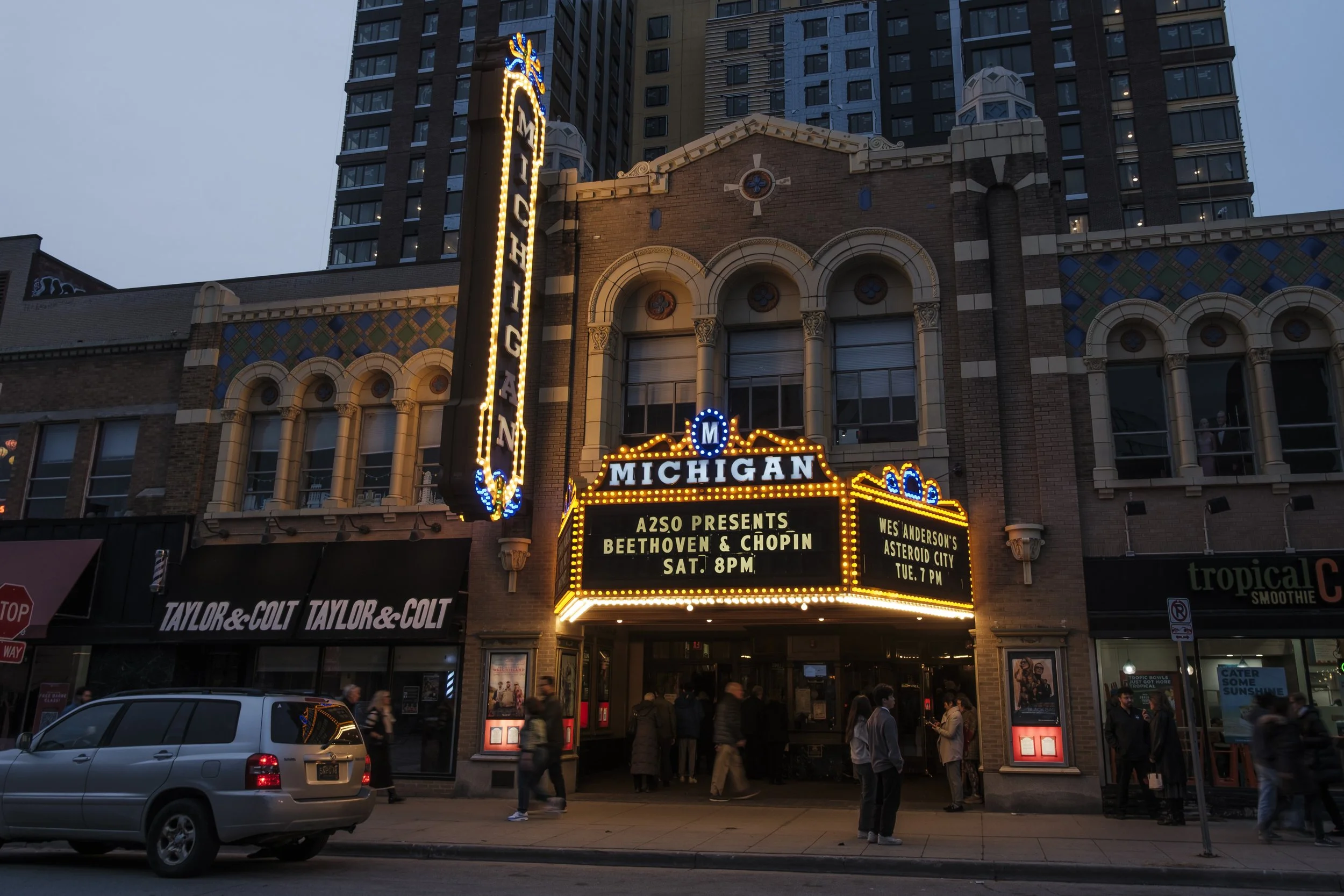 The Michigan Theater in downtown ann arbor is illuminated by a marquee while blurry people walk on the sidewalk.