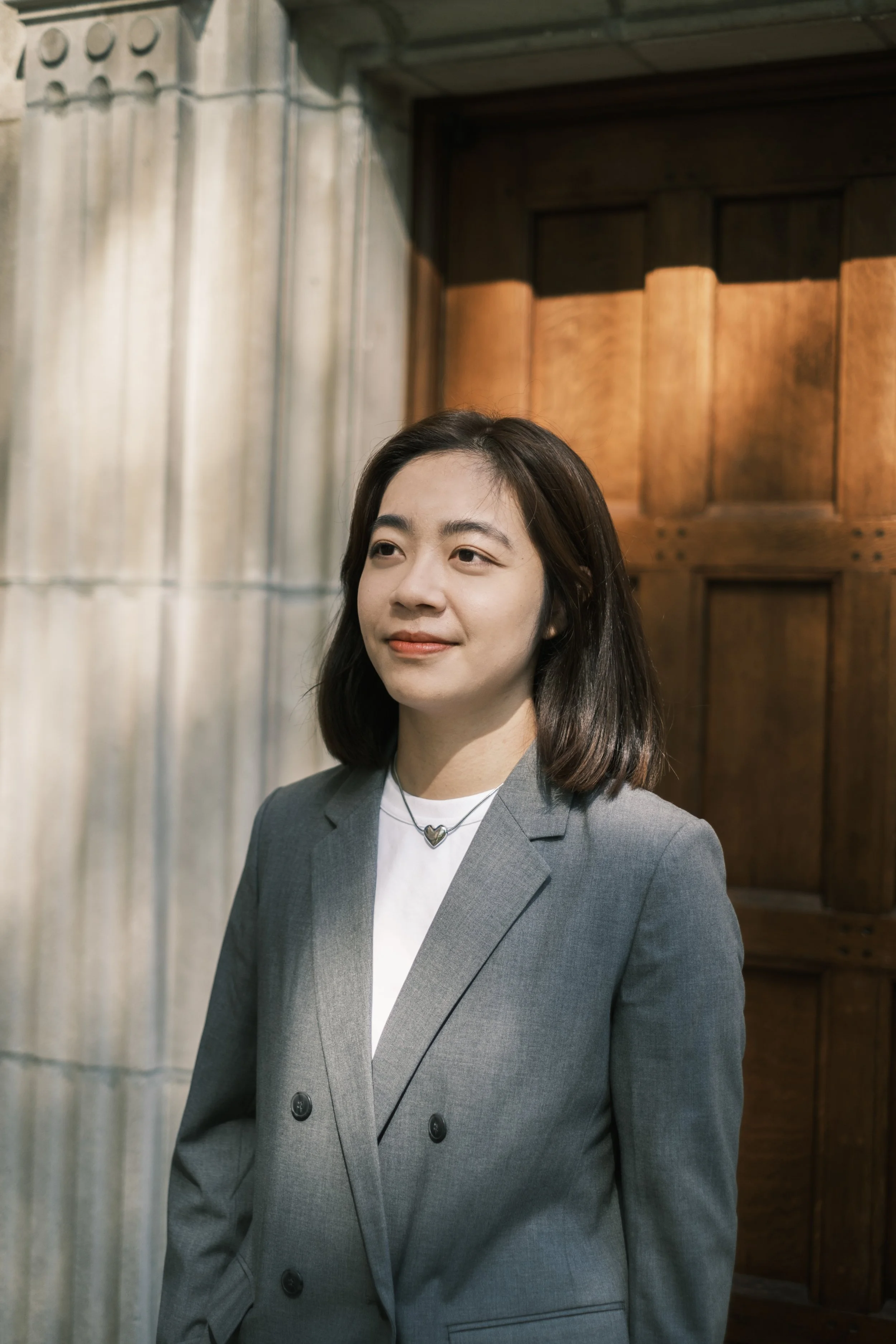 A woman stands wearing a gray suit and a heart necklace while standing near a doorway.