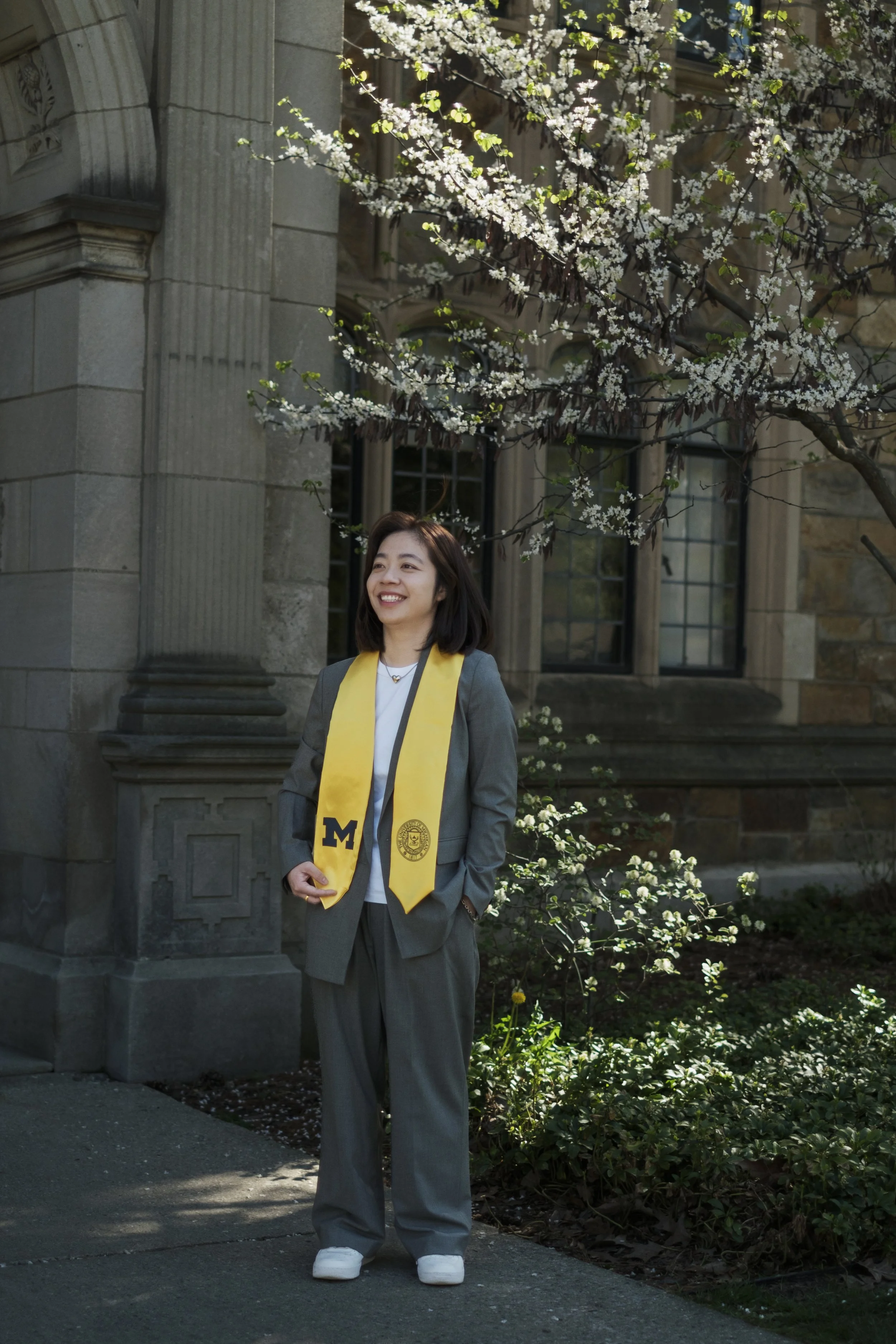 A woman stands, smiling, wearing a university of michigan graduation sash in front of trees and a brick building.
