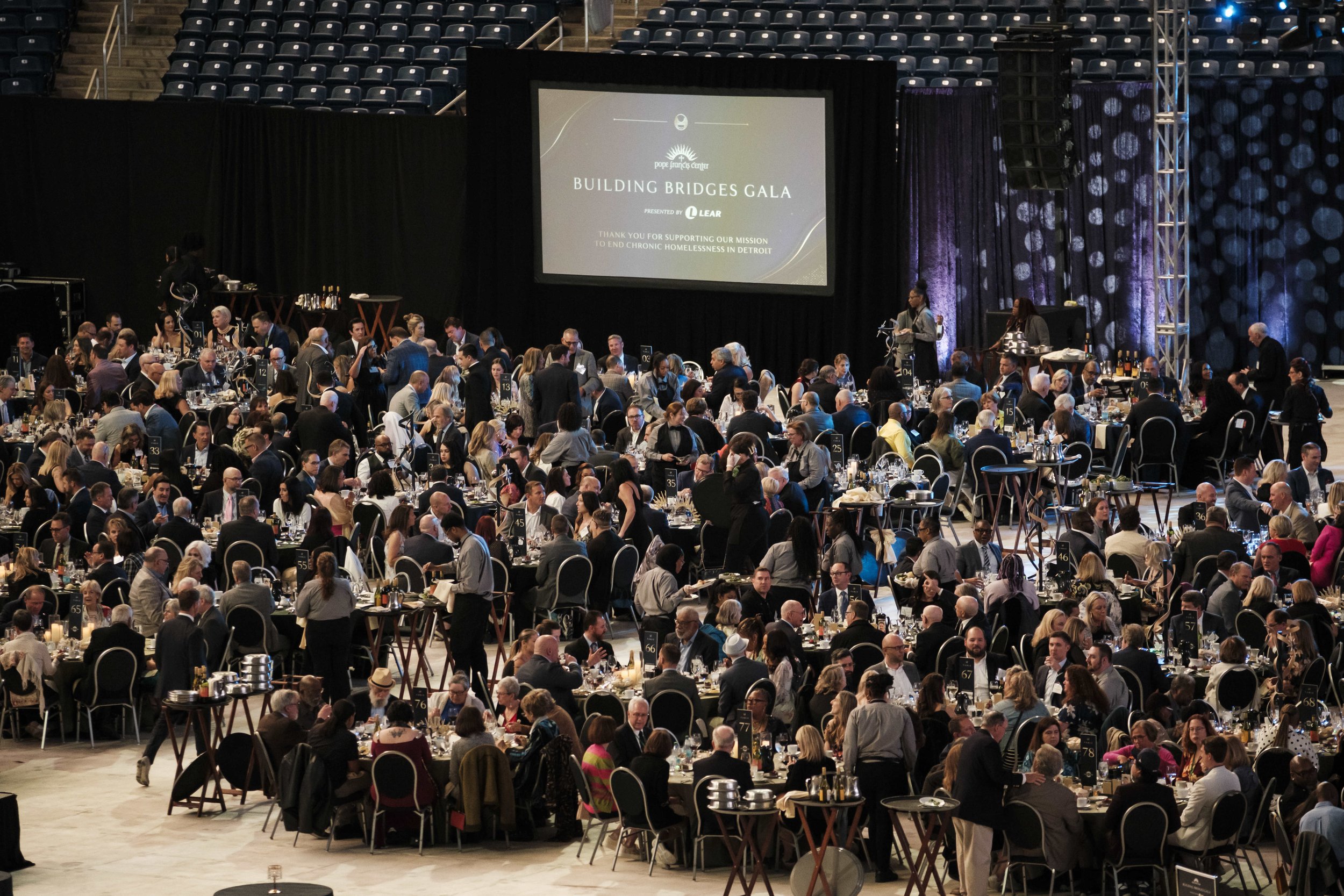 A high up view of people among tables during an event with large screen that says "Building Bridges Gala"