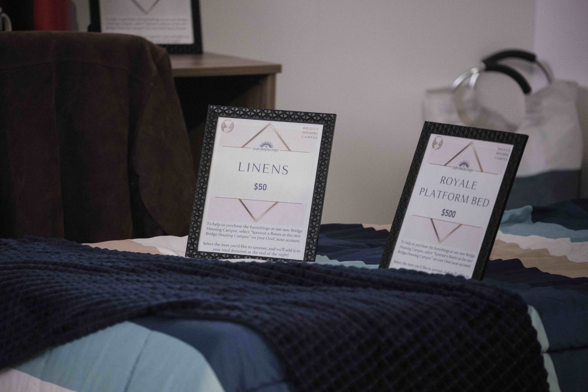 Framed signs laying on a bed that list bedding items and the cost for each item.