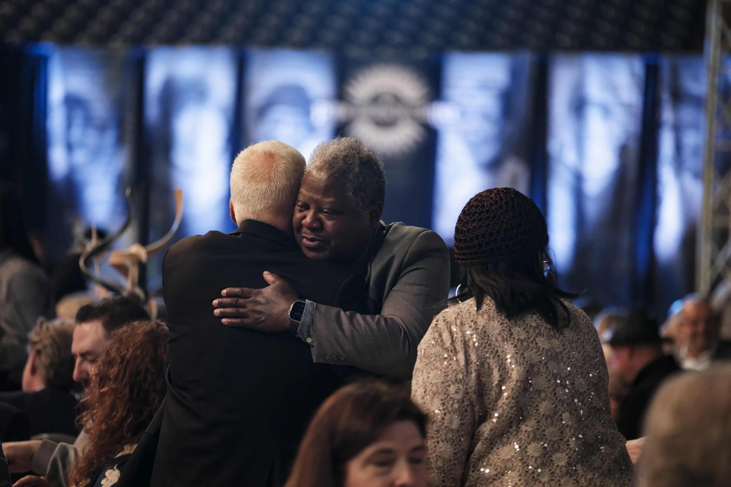Two men hug in a crowd of people against a blue background.